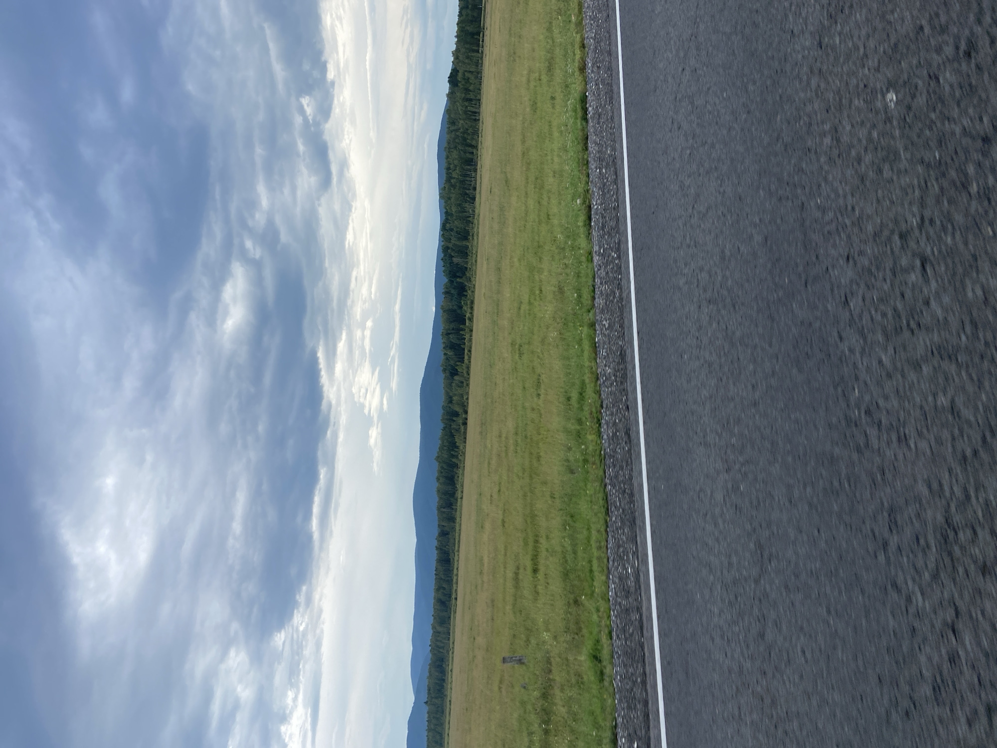 An overcast day in Zun-Murino, Russia, as seen from a roadside. The foreground features a dark asphalt road with a distinct white line marking the lane. Beyond the road lies a vast, flat expanse of green grassland dotted with small wildflowers and a single wooden post. This field stretches towards a dense, dark green forest that forms a clear horizontal line across the middle ground. Behind the forest, layered, hazy blue mountains rise against the sky. The sky itself is filled with dramatic, heavy grey clouds, with patches of lighter grey and white indicating breaks or thinner cloud cover. The overall scene evokes a sense of natural, open space under a moody sky.