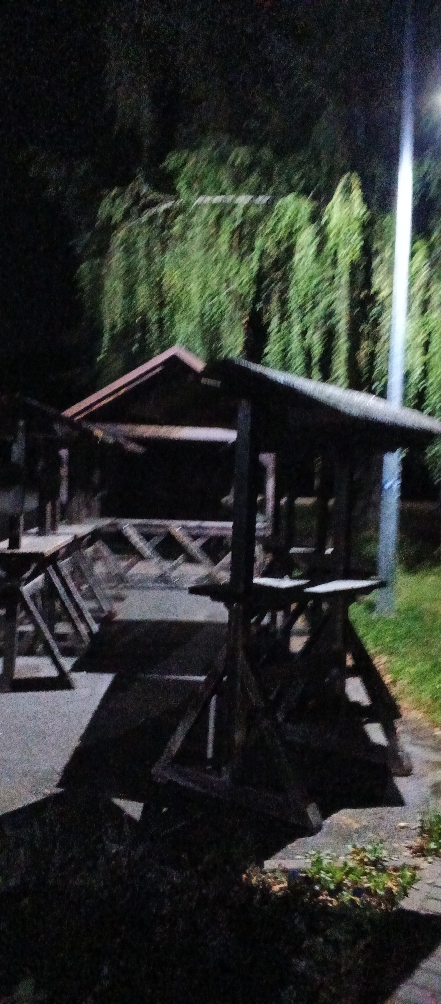 An outdoor scene at night, with a wooden structure resembling a market stall or pavilion in the foreground. The structure has a peaked roof and is supported by wooden posts. There are tables or benches within the structure. In the background, there are trees with drooping branches, illuminated by a light source, possibly a streetlamp, visible to the right. The ground appears to be paved with some patches of grass and foliage. The image is blurry and lacks detail due to low light conditions. There are no people visible in the image. The scene does not contain any identifiable landmarks or features that would suggest a specific location within the city of Mukachevo, Ukraine.