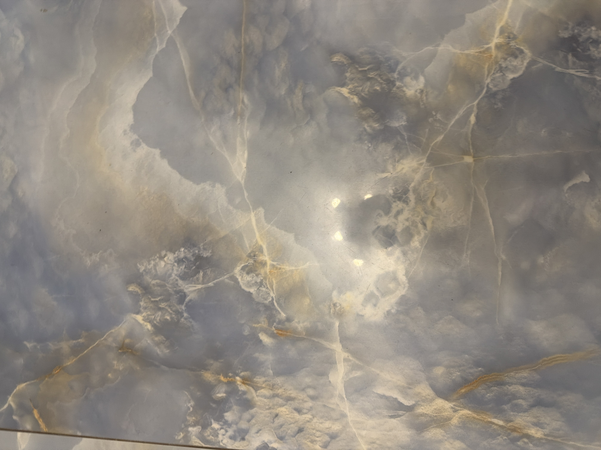 An overhead shot shows a marble tile with a marbled blue and white pattern. The tile is veined with thin lines of golden brown and white, creating a swirling, cloud-like effect. There are some bright reflections on the surface of the tile, suggesting it is polished. The tile appears to be part of a larger surface, possibly a floor or countertop, as a thin vertical line is visible on the far left edge, indicating a grout line.