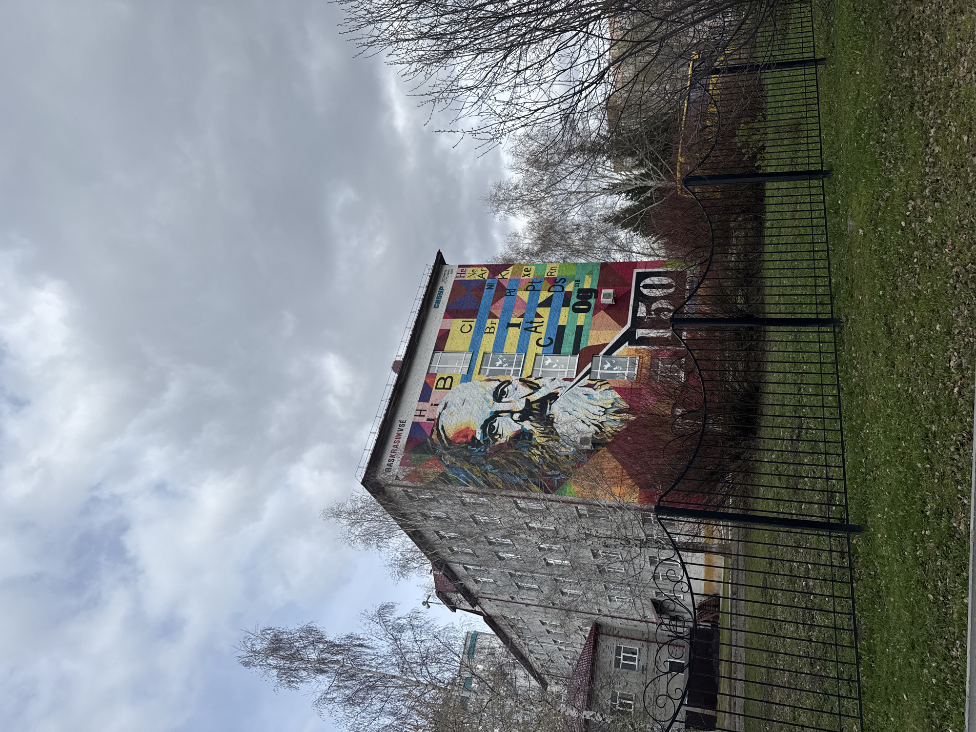 The image shows a tall, multi-story building in Tobolsk, Russia, featuring a large mural of Dmitri Mendeleev, the creator of the periodic table, on its side. The mural also includes a colorful representation of the periodic table itself. The building's exterior appears to be primarily gray and textured. In the foreground, there's a black metal fence running parallel to the building. Beyond the fence, there's a strip of green grass and bare trees in the background, suggesting it might be late fall or early spring. The sky is overcast with gray clouds, creating a muted, cold atmosphere.