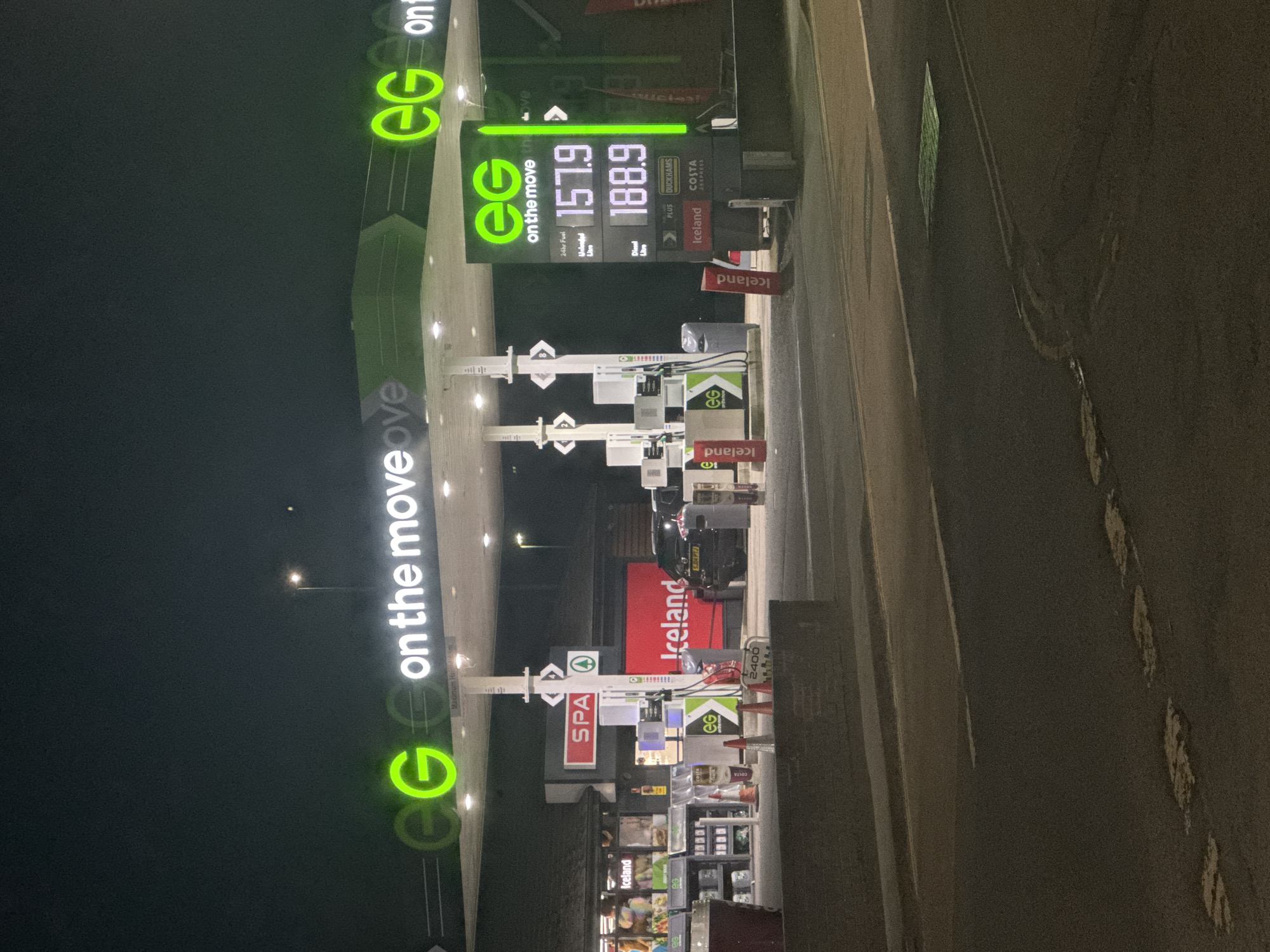 The image depicts an EG petrol station, located in Welshpool, United Kingdom. The foreground is illuminated by the station's lights, showcasing fuel prices: 157.9 for unleaded fuel and 188.9 for diesel per litre. Several fuel pumps are visible under illuminated canopies with EG branding. In the mid-ground, a convenience store is identifiable by Iceland branding along with advertising for Costa Express. The background is dark, suggesting night. A car is parked near the pumps. The scene includes signage for SPA and Duckhams motor oil.