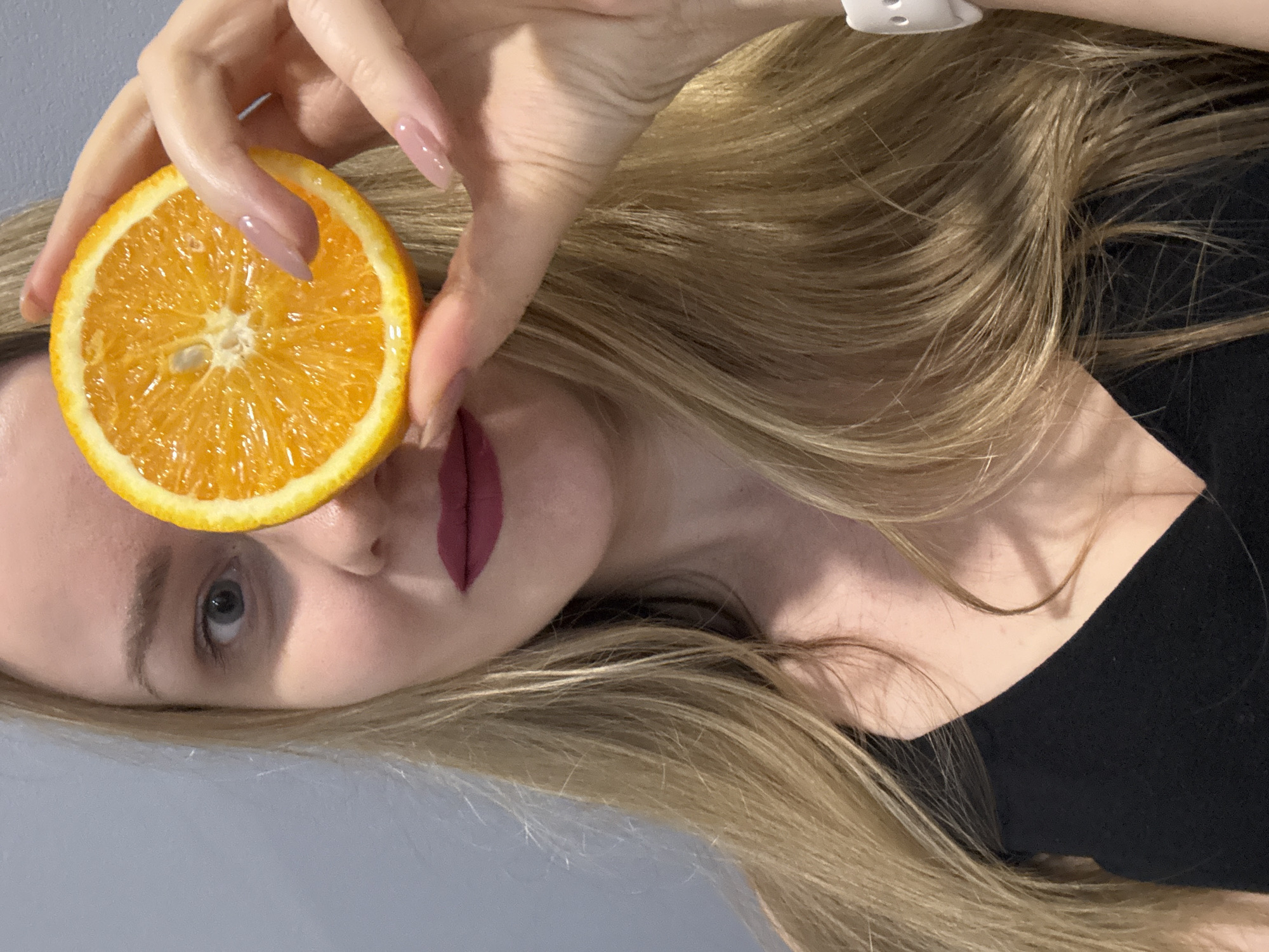 A fair-skinned woman with blonde hair is holding a slice of orange over her left eye. She has mauve lipstick on, a light eye color, and light pink manicured nails. The background is a light gray color. The woman is wearing a black top. There is no indication of the location as the city of Chaikovsky, Russia.