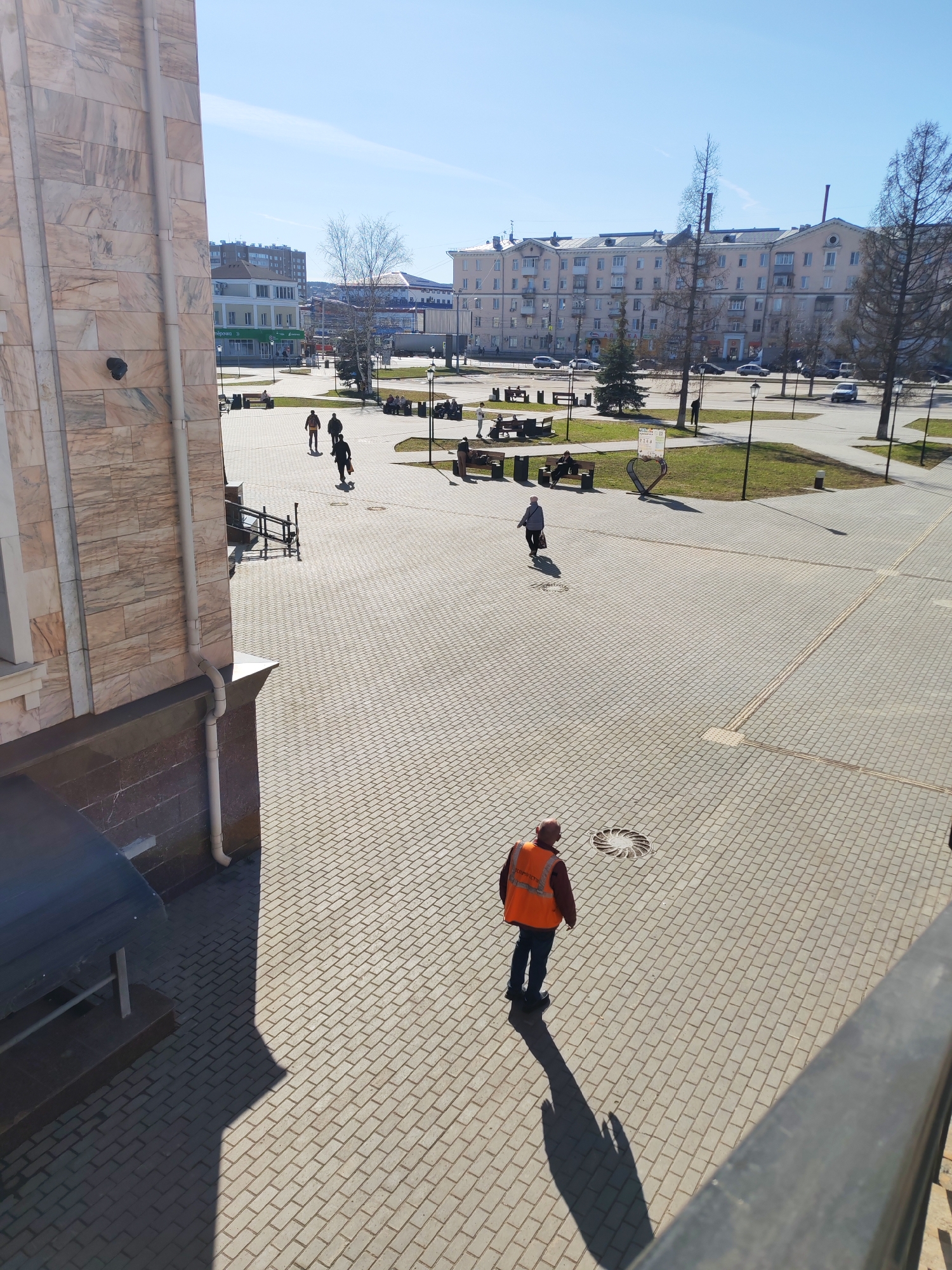 The image presents an elevated view of a paved city square or pedestrian area in Izhevsk, Russia, under a clear, bright sky. The ground is extensively covered with light-colored, rectangular pavers laid in a basketweave pattern.
In the immediate foreground, facing away from the viewer, stands a man wearing a bright orange safety vest over a dark jacket and blue trousers. A partial Russian inscription, possibly "КОММУНАЛЬНЫЕ" (Communal Services), is visible on his vest. His long shadow extends forward, indicating a low sun angle, characteristic of morning or late afternoon. A circular manhole cover with a decorative spiral pattern is visible near his feet.
Further into the scene, several other individuals are present; some walk along the paved paths, while others are seated on benches located within grassy areas. Bare trees suggest late winter or early spring. A building with light-colored, possibly marble or granite, cladding dominates the left side of the frame, featuring a visible rain gutter pipe. In the background, a street with parked and moving cars separates the square from more distant urban buildings, including one with a visible "ПЯТЁРОЧКА" (Pyaterochka) supermarket sign. Other architectural styles, typical of a Russian city, are also present. The square is adorned with street lamps and a distinctive heart-shaped metal sculpture or sign on a grassy patch. The overall scene depicts everyday activity in a sunny urban public space.