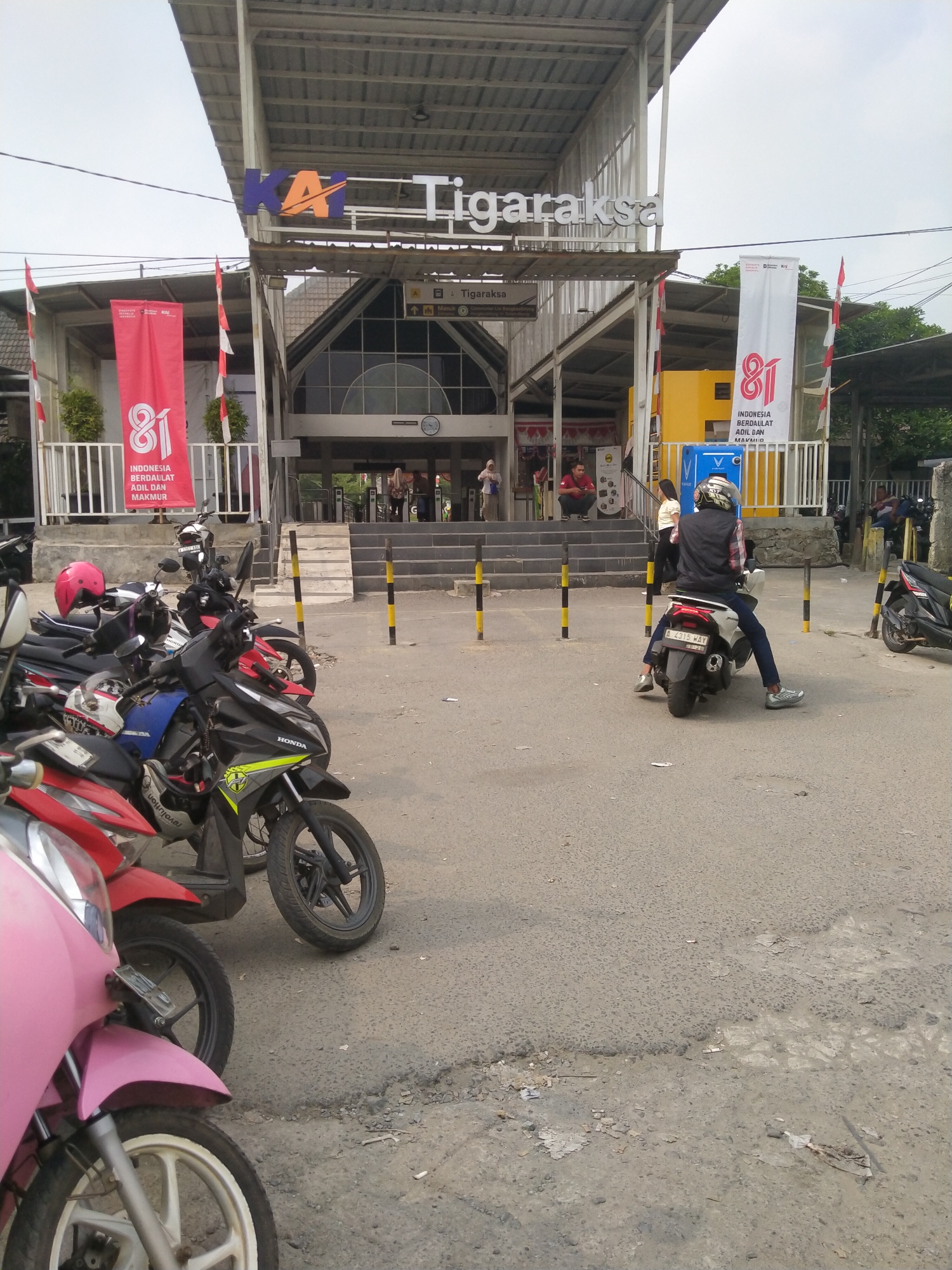 This image depicts the entrance to the Tigaraksa train station in Cikuya, Indonesia. In the foreground, several motorcycles are parked on a paved area. One pink scooter is partially visible on the left. Behind the parked motorcycles, a person is seated on a white scooter, wearing a helmet, vest, and dark pants. They are facing away from the camera, looking towards the station. The station itself features a prominent KAI logo and "Tigaraksa" signage above the entrance. Red and white banners, likely celebrating Indonesia's 81st Independence Day, are displayed on either side of the entrance. People can be seen on the steps leading into the station, some standing and others sitting. The environment appears to be an overcast day, with a mix of paved ground and a worn asphalt surface with some debris.