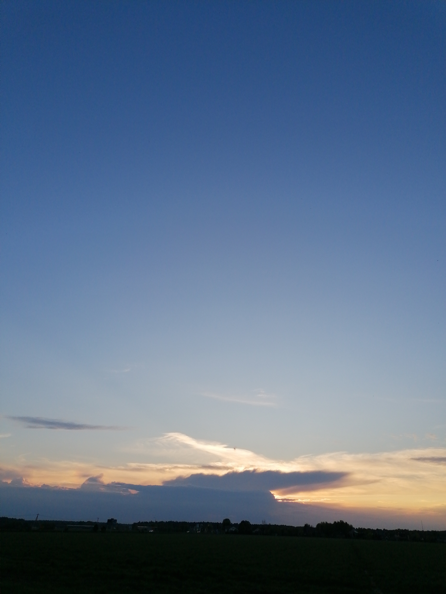 The image shows a landscape at sunset. The sky transitions from blue above to a warm, golden hue near the horizon. Several layers of clouds are present, some dark and opaque, others translucent and reflecting the sunlight. The horizon line is comprised of a dark, silhouetted landscape, likely fields and trees. There are no people visible in the image. The location is unknown.