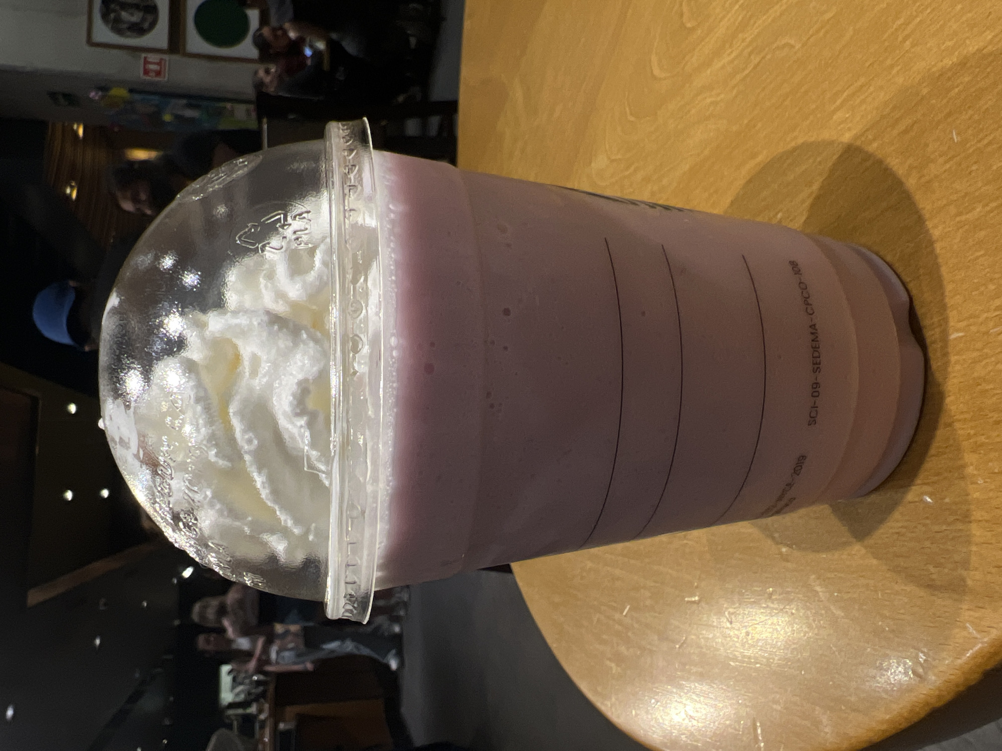 A Starbucks Frappuccino is displayed on a tan wooden table in Mexico City, Mexico. The beverage is a light purple smoothie with whipped cream on top, sealed with a clear plastic dome lid. The Starbucks cup has markings including "SCI-09-SEDEMA-CPCO-108." The background shows a dimly lit interior with other patrons, reflecting in the plastic lid.