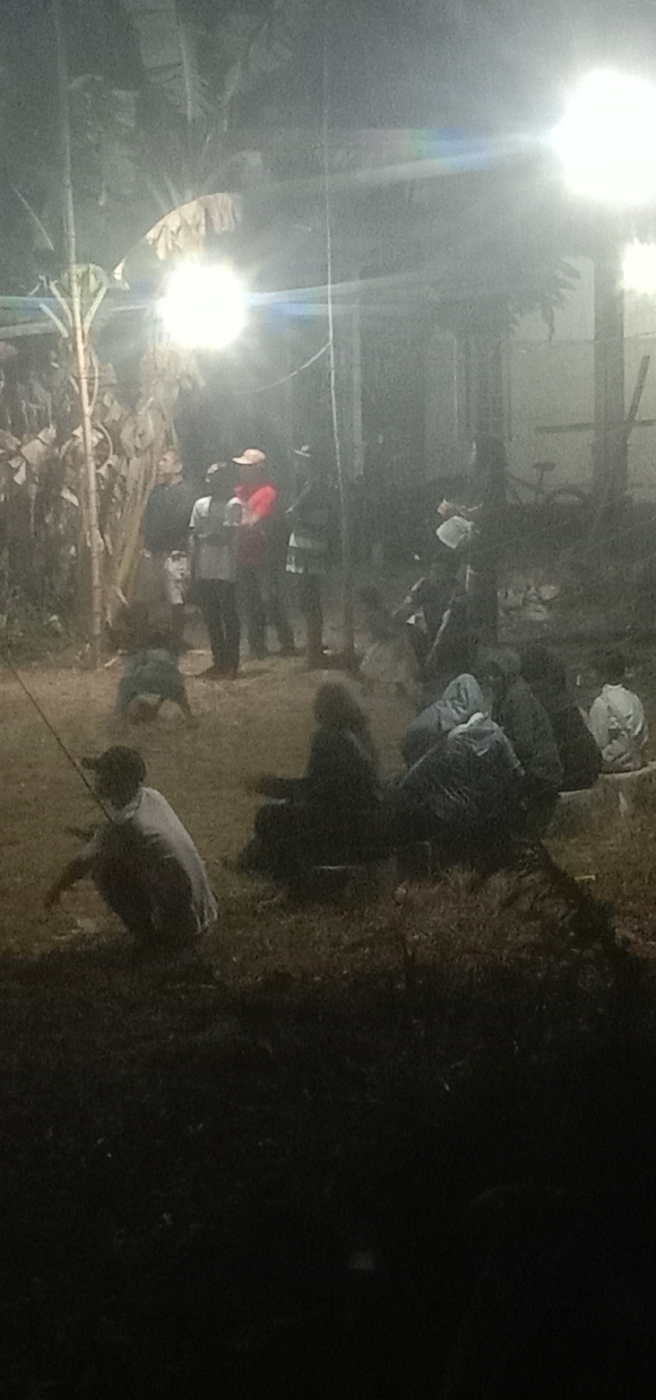 An outdoor scene at night in Banjar Dowo, Indonesia, is illuminated by artificial lights, casting a haze over the environment. A group of people are gathered, some standing and others sitting or crouching. In the foreground, a person is crouched down, holding a thin pole that extends to the left. To their right, other individuals are seated on the ground, some with hoods pulled up. In the midground, a cluster of people are standing, one person wearing a red jacket and a light-colored cap. Behind them, more figures are visible. The ground appears to be a mix of dirt and grass, with vegetation, including what look like banana trees, present in the background. The overall impression is of a casual gathering or event taking place in a somewhat rustic, outdoor setting.