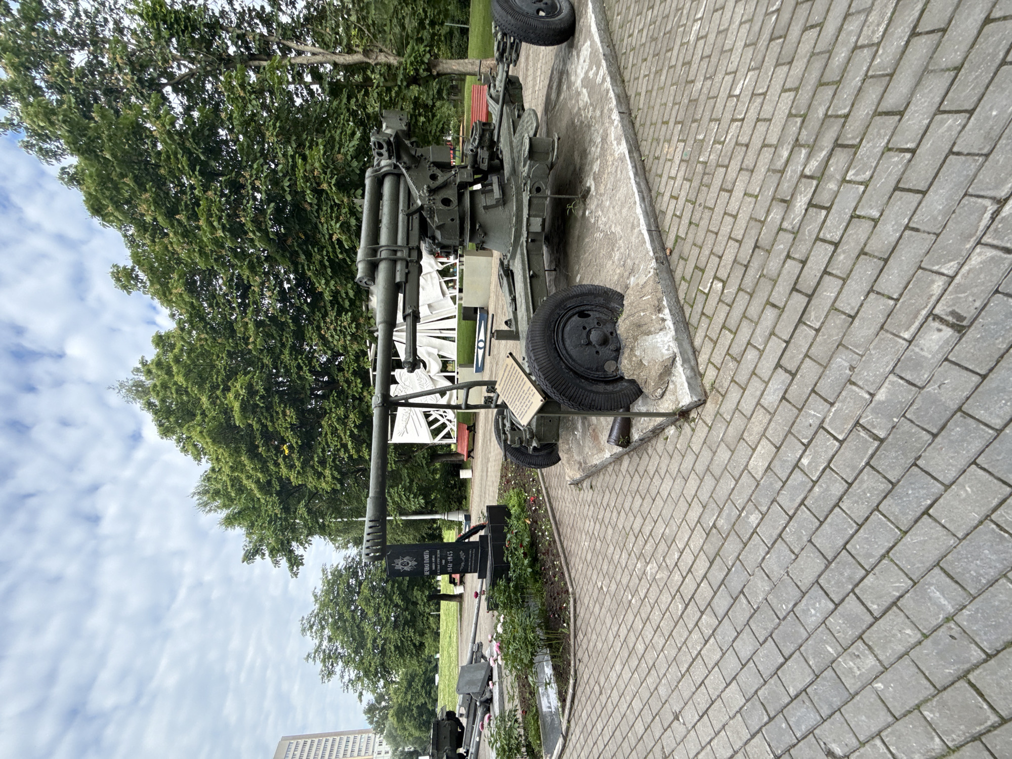 The image depicts a war memorial in Moscow, Russia. In the foreground, an anti-aircraft gun is displayed on a concrete base, with two large wheels visible. A plaque with text is affixed to the front of the gun. Behind the gun, a black granite obelisk stands, bearing an inscription and a wreath. Flowerbeds with pink and white flowers surround the base of the obelisk. In the background, mature green trees form a dense canopy, and a tall, modern building is visible on the left. The ground is paved with gray bricks. The sky is cloudy, with wispy white clouds against a pale blue background.