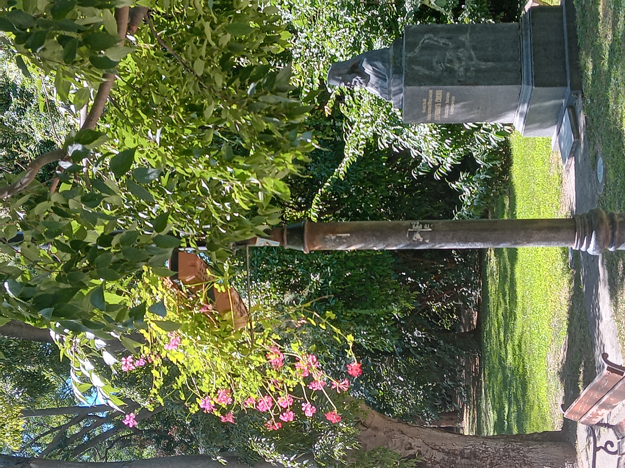 This is a photograph of a park in Varna, Bulgaria, featuring a bust of a historical figure on a pedestal, a lamp post, and a bench. The bust, made of dark stone, depicts a man with a beard and a head covering, likely a religious or historical figure. The pedestal beneath it is inscribed with text, though it is partially obscured. A weathered, dark-colored lamp post stands to the left of the bust, closer to the foreground. A wooden park bench with decorative metal legs is visible in the lower left corner. Lush green trees and foliage dominate the background, with dappled sunlight filtering through the leaves, suggesting a daytime scene. Pink flowers are blooming on vines in the upper left portion of the image. The ground is covered with grass and a paved path. The overall setting is a well-maintained public park.