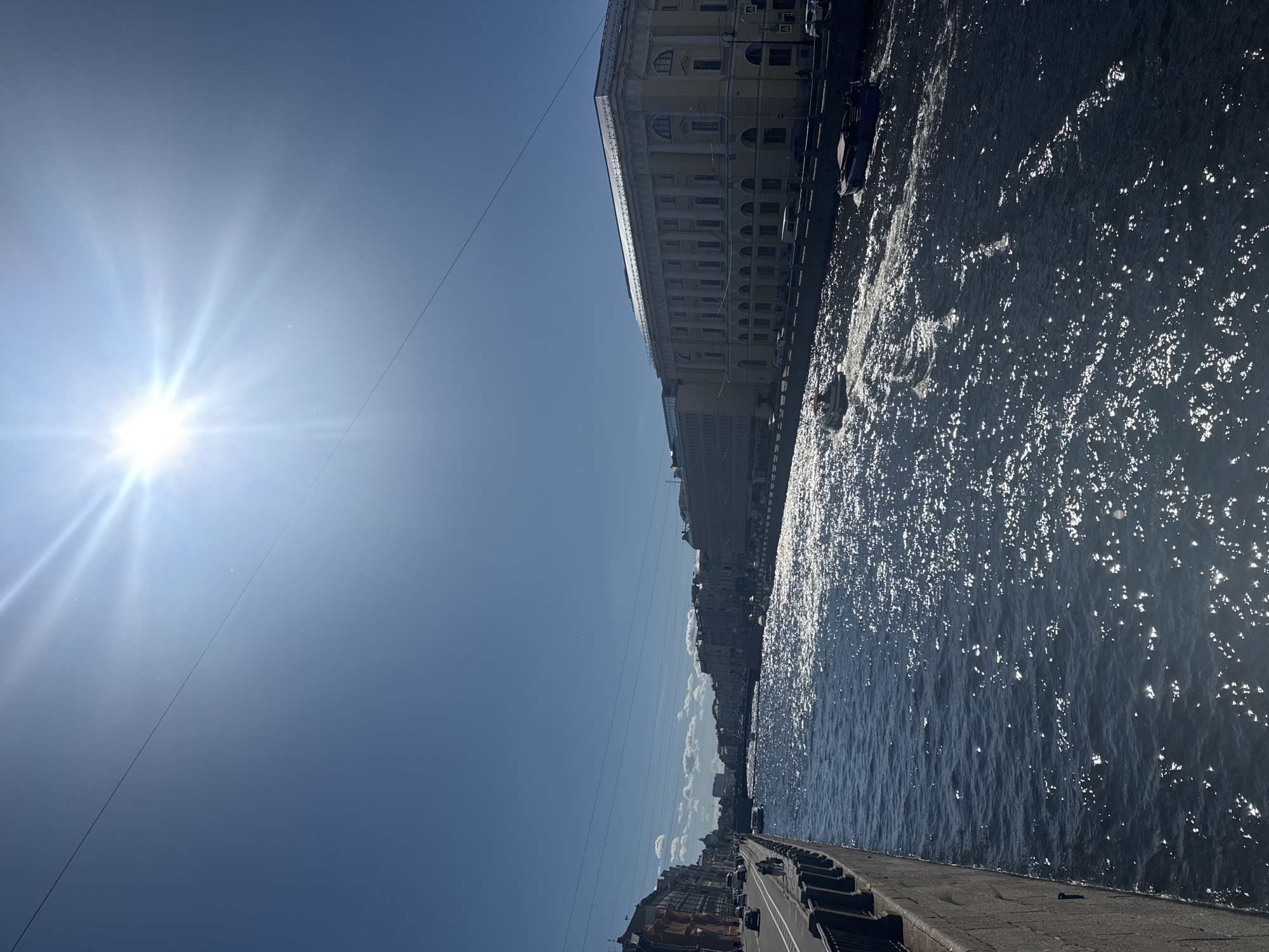 The image depicts a sunny day on a canal in Saint Petersburg, Russia. The sun is shining brightly, creating a glare on the water. On the left side of the canal, there is a paved walkway with a black railing, and beyond that, buildings line the street. Cars are visible on the street. On the right side of the canal, there are several large, classical-style buildings with columns. Two boats are in the canal, one white and one dark blue, both moving and leaving wakes. The sky is clear blue with a few wispy clouds in the distance. The water of the canal is dark blue and appears choppy due to the sunlight reflecting off its surface and the movement of the boats.