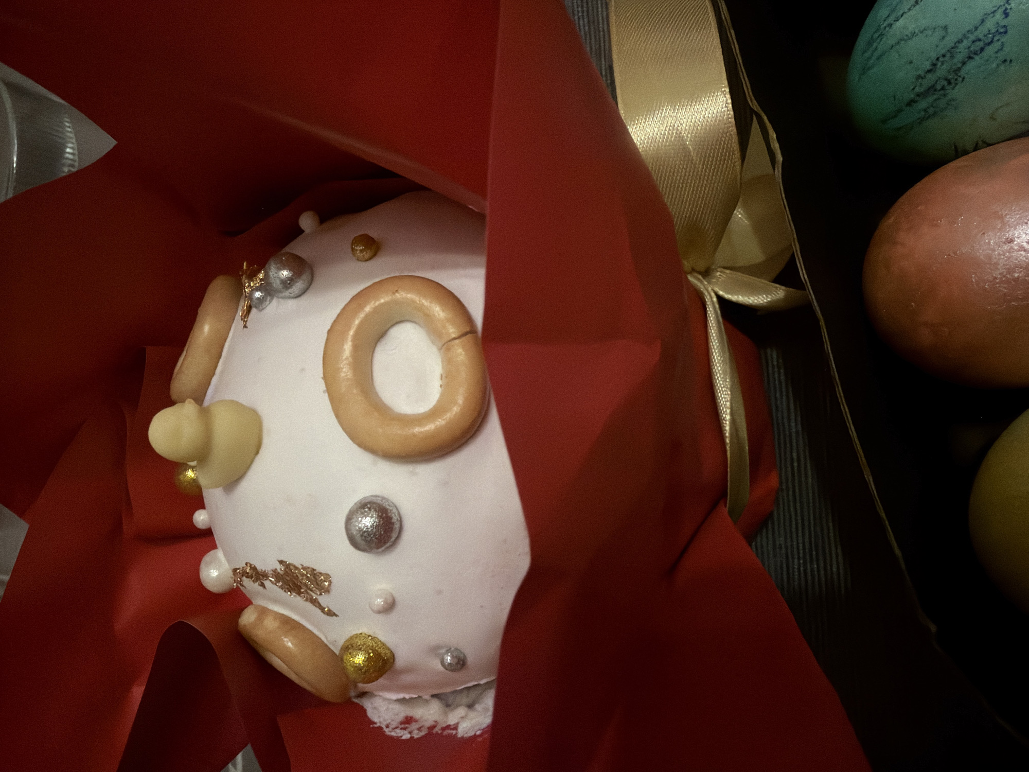 A close-up shot depicts a decorated Easter cake, likely a Kulich, wrapped in red paper. The cake is adorned with white icing and decorated with small bagels, silver and gold beads, and what appear to be white chocolate candies. Beside the cake, there are some painted Easter eggs. The presence of Kulich suggests that the location is likely Moscow, Russia, or a place with a significant Russian Orthodox population, as Kulich is a traditional Easter bread in that culture.