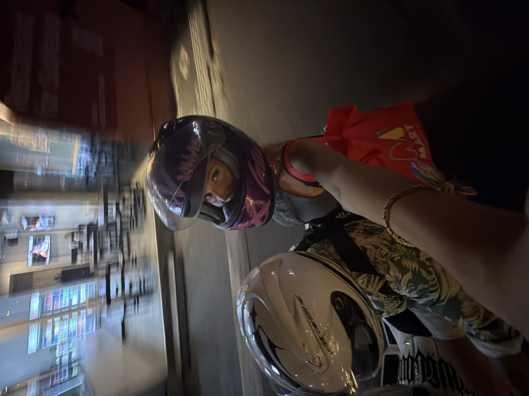 This image captures a perspective from the back of a motorcycle as it travels down a street in North Kuta, Indonesia. The driver, wearing a white helmet with black and blue markings and the text "WE CAN STOP ME" visible, is in the foreground. Behind them, a passenger wearing a purple helmet with pink accents is looking towards the camera. They are wearing a dark tank top and have a red bag with a "MART" logo hanging from their shoulder. The background is blurred due to motion, showing a street scene with what appears to be a storefront or restaurant with tables and chairs inside. The lighting suggests it is nighttime or dusk.