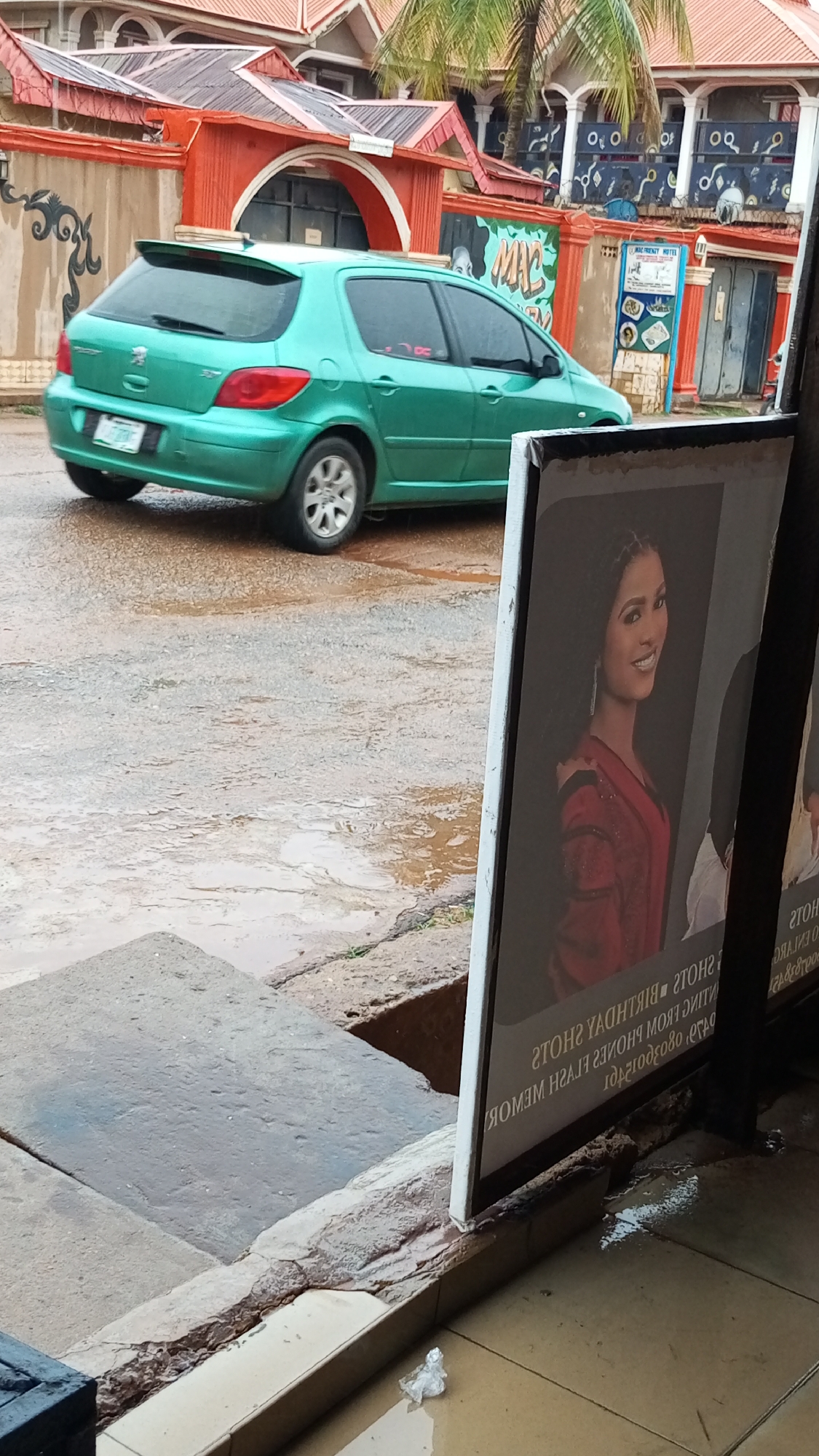 The scene depicts a street in Sabon Tasha, Nigeria, with a green Peugeot hatchback driving on a wet road. A promotional sign is in the foreground displaying a woman's portrait. The road shows signs of recent rainfall. A building with a red roof is visible in the background.