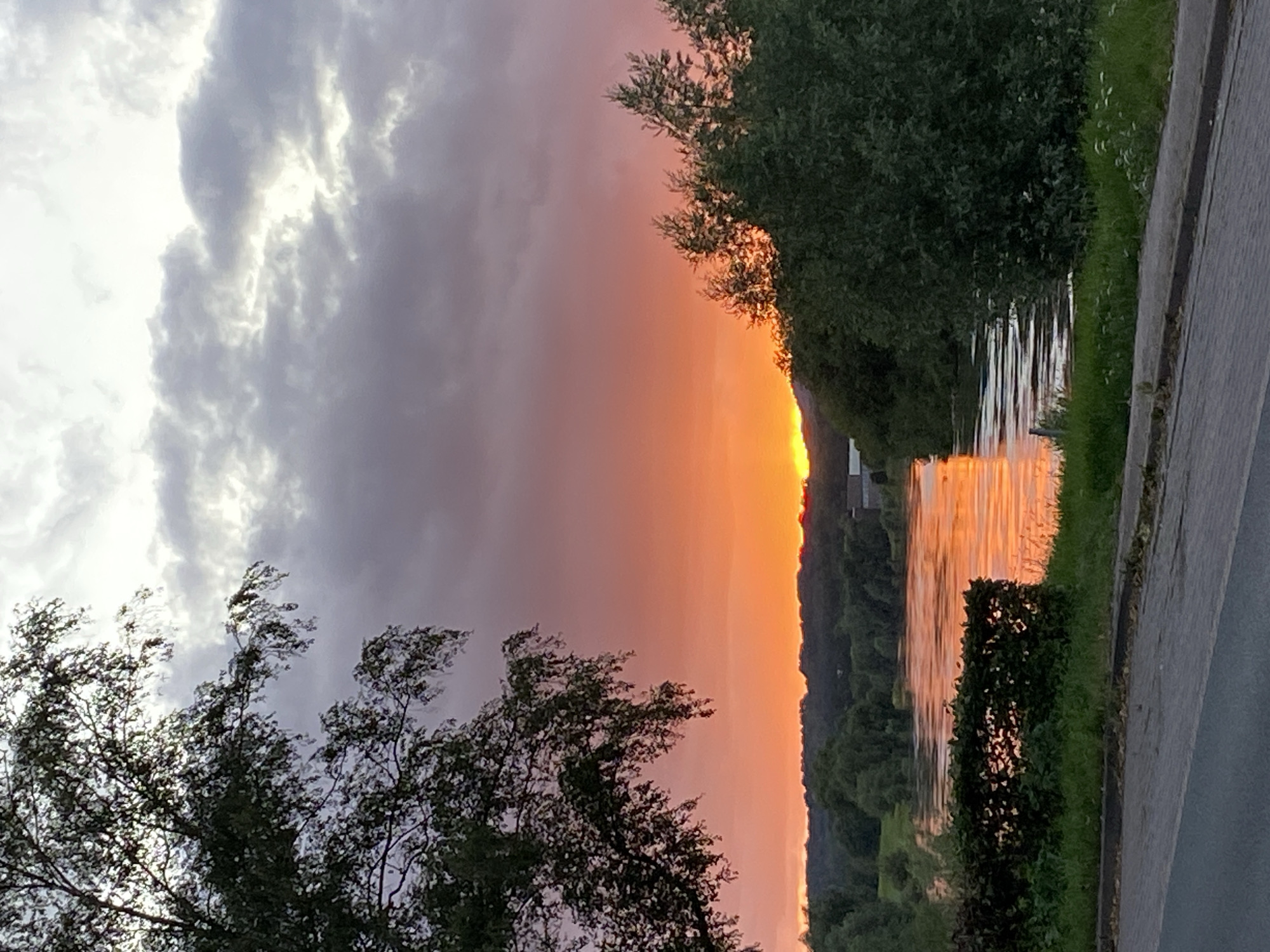 An image captured at sunset in Rinteln, Germany, shows a wide river reflecting the vibrant orange and pink hues of the sky. The foreground features a paved road and a manicured hedge. Lush green trees and foliage line the riverbanks, with a dense forest forming the backdrop under a sky filled with dramatic, textured clouds. The setting sun is partially obscured by the distant treeline, casting a warm glow across the water and surrounding landscape.
