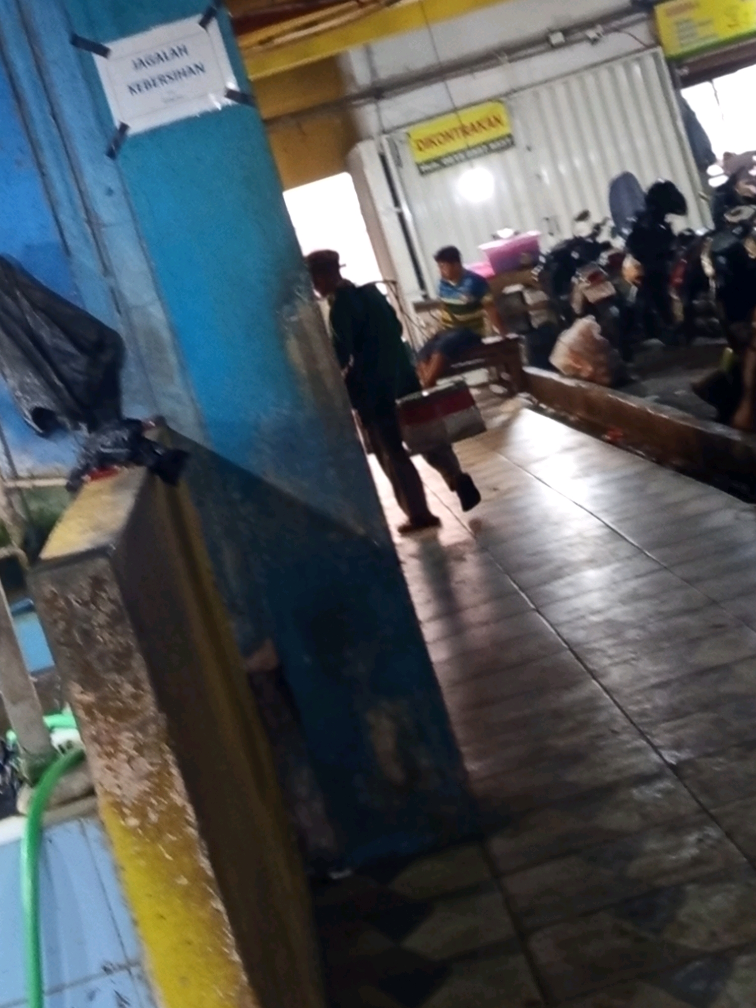 The image shows a covered outdoor area, possibly a parking lot or a market stall, with a tiled floor. There are several motorcycles parked in the background. A person wearing a dark shirt and a hat is walking away from the camera, carrying a red and white object. Another person is sitting in the background. A sign is visible on a blue pillar, which appears to read "JAGALAH KEBERSIHAN" (Maintain Cleanliness). Another yellow sign in the background states "DIKONTRAKAN" (For Rent) and provides a phone number. The scene suggests a commercial or public space in Karawang, Indonesia, with an emphasis on cleanliness.
