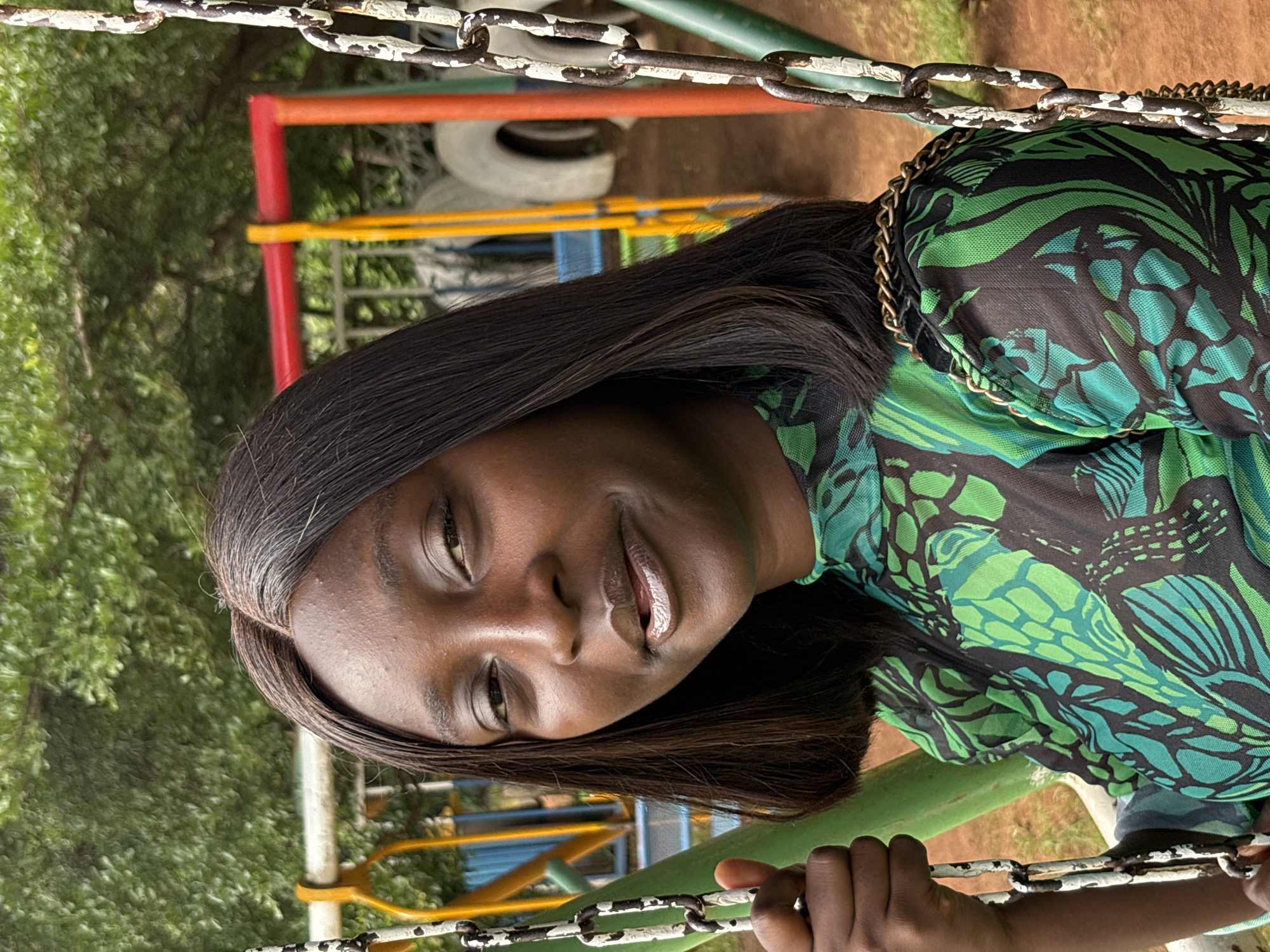 A portrait of a young Black woman is shown, with the image rotated 90 degrees counter-clockwise. She is positioned in the foreground, with her head tilted and looking downwards towards the viewer. Her dark, straight hair frames her face, and she wears a green and black patterned top with a gold chain detail around the neckline. Her lips are slightly parted and appear to have a light-colored lip gloss.
In the background, a playground structure is visible, with red, yellow, and green bars. A tire is also present behind the structure. The setting appears to be outdoors, with lush green foliage suggesting a natural, possibly park-like environment. The lighting suggests it is daytime, with no specific weather conditions being evident. The overall mood is casual and relaxed. The location is identified as Bukuru, Nigeria.