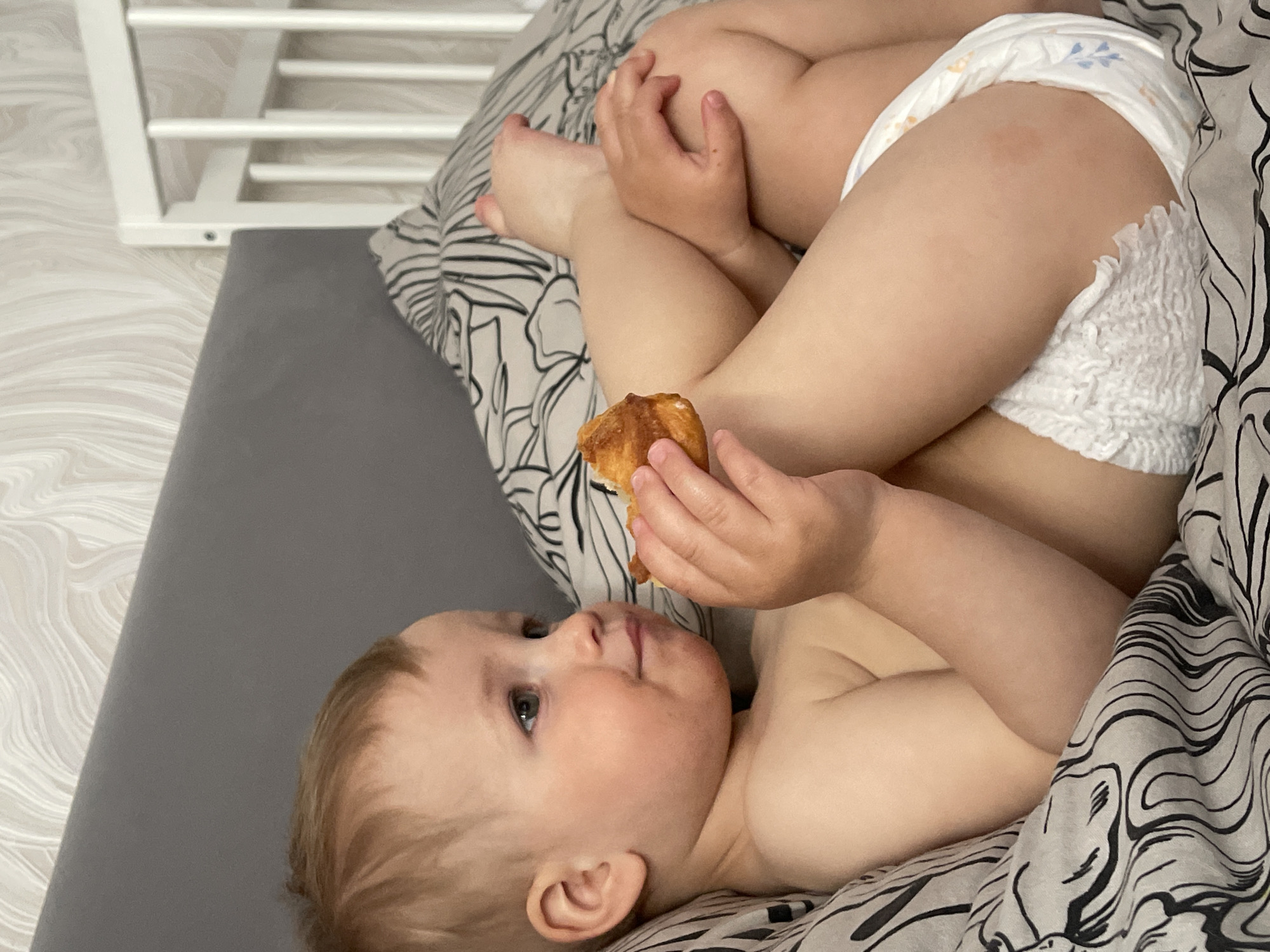 A fair-skinned, blond toddler wearing a diaper is lying on a bed covered with a black and white patterned blanket. The toddler is holding a piece of golden-brown pastry in its hands and looking to the left. To the left of the toddler is a gray surface and a white structure resembling a crib or a piece of furniture. The image appears to be taken indoors. There is no indication of Izhevsk, Russia.