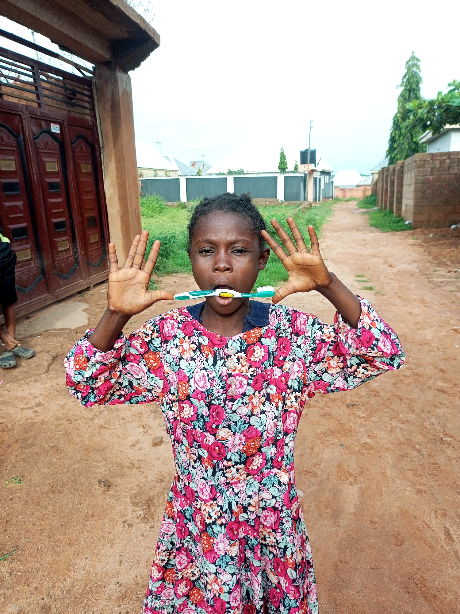 A young girl with dark skin and braided hair is standing outside, holding a toothbrush horizontally in her mouth. She is wearing a colorful floral dress with a navy blue collar. Her arms are raised with her palms facing outwards. Behind her, there is a dirt road, a brick wall, some greenery, and buildings. The environment suggests a semi-urban or rural setting in Tempure, Nigeria, possibly a residential area. The lighting indicates it is daytime.
