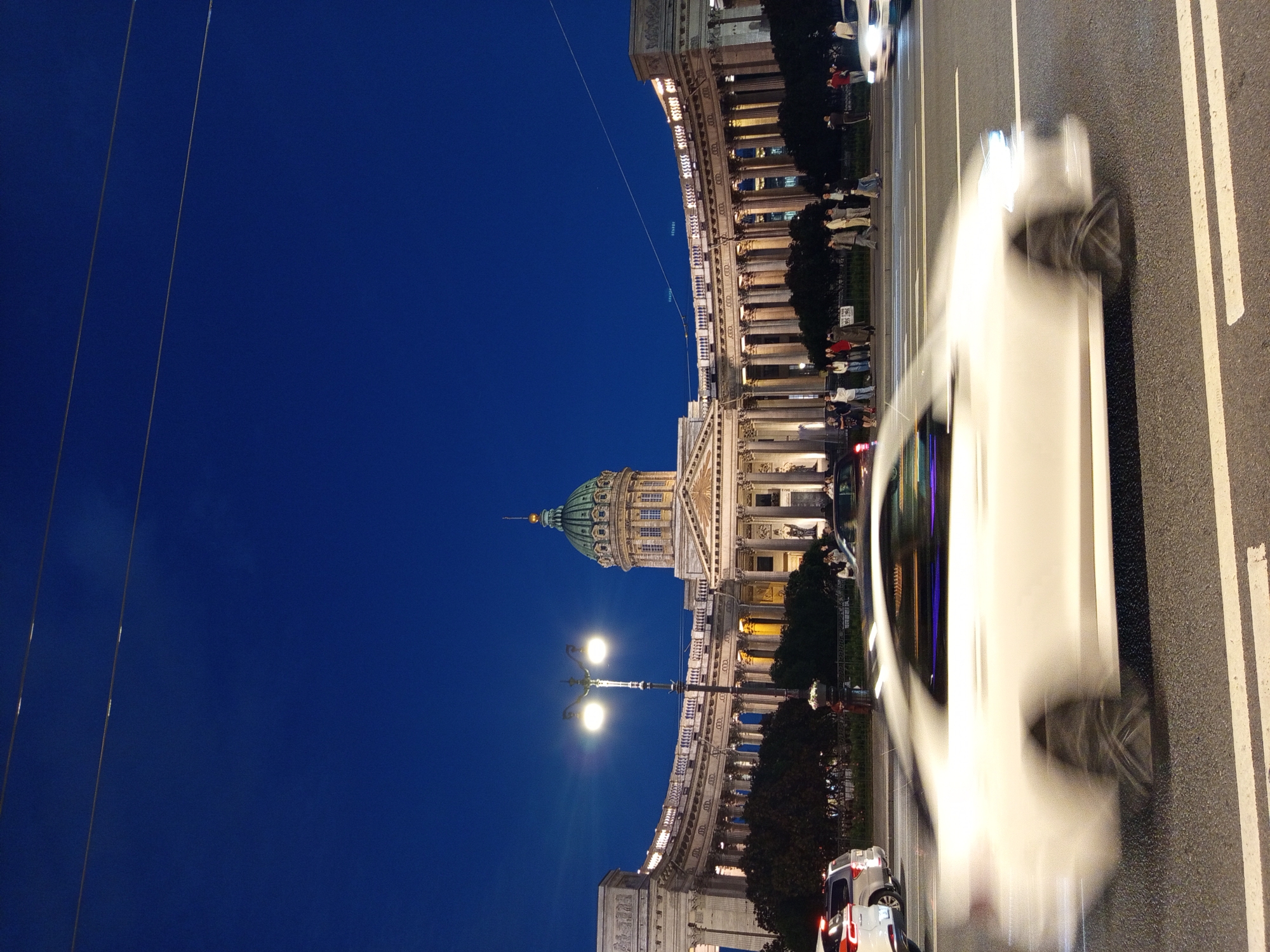 This is a full-frame, vertical, eye-level shot of Kazan Cathedral at dusk in Saint Petersburg, Russia. The facade of the cathedral is illuminated by external lighting, with its large, green dome prominently visible against the deep blue twilight sky. In the foreground, a white car is captured in motion blur, indicating a slow shutter speed. Several other vehicles and a lamppost are also present on the street. A group of pedestrians is visible on the sidewalk in front of the cathedral, some of whom are facing the building and appear to be observing it. The overall scene depicts a bustling urban environment with a significant architectural landmark.