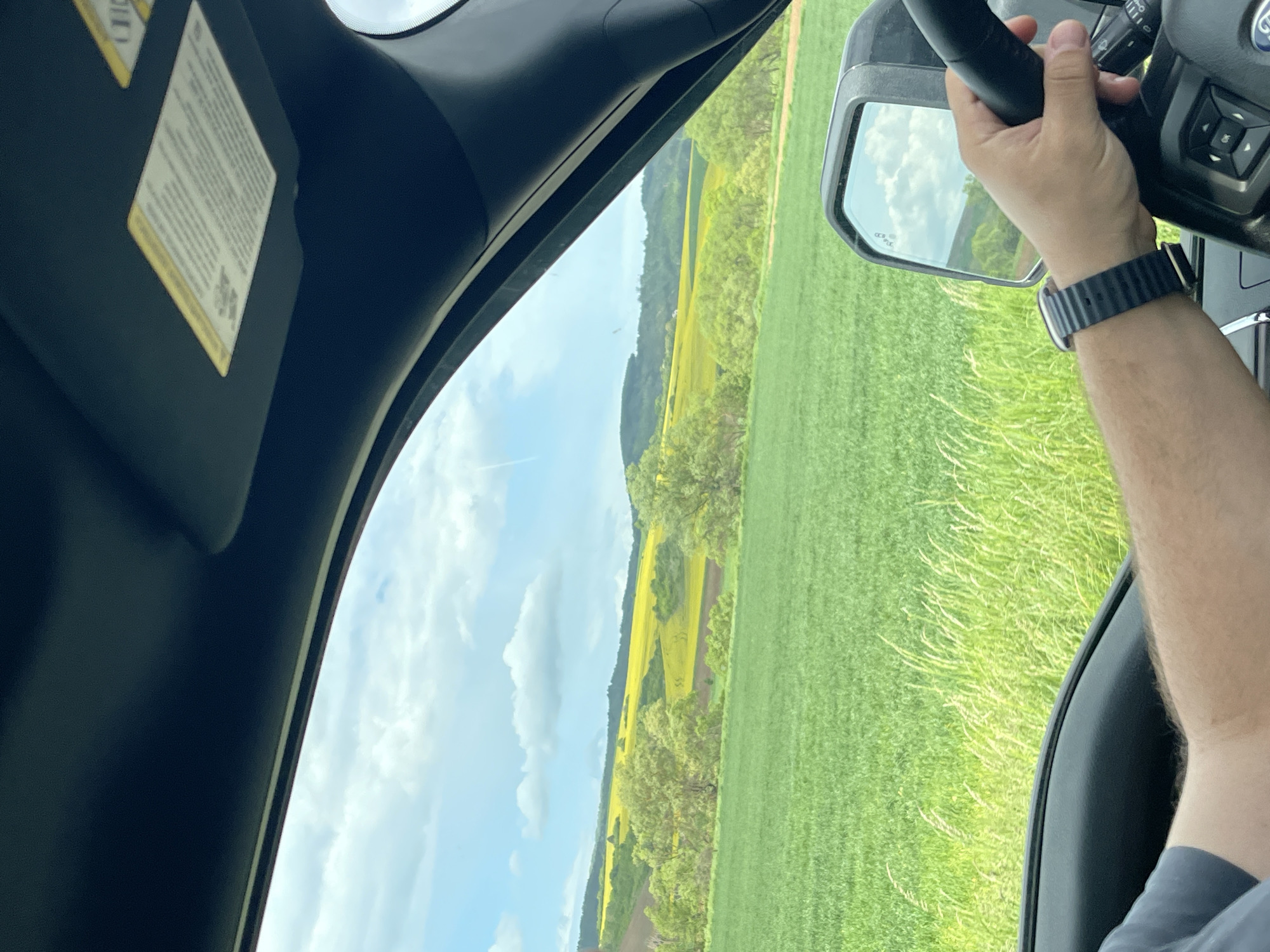 A car is driving through a green field landscape. The driver's hand, wearing a watch, is visible on the steering wheel, and the side mirror reflects the scenery. The landscape includes a line of yellow crops or flowers and a partly cloudy sky. The location, Yardzhilovtsi, Bulgaria, is not visually discernible from this image.