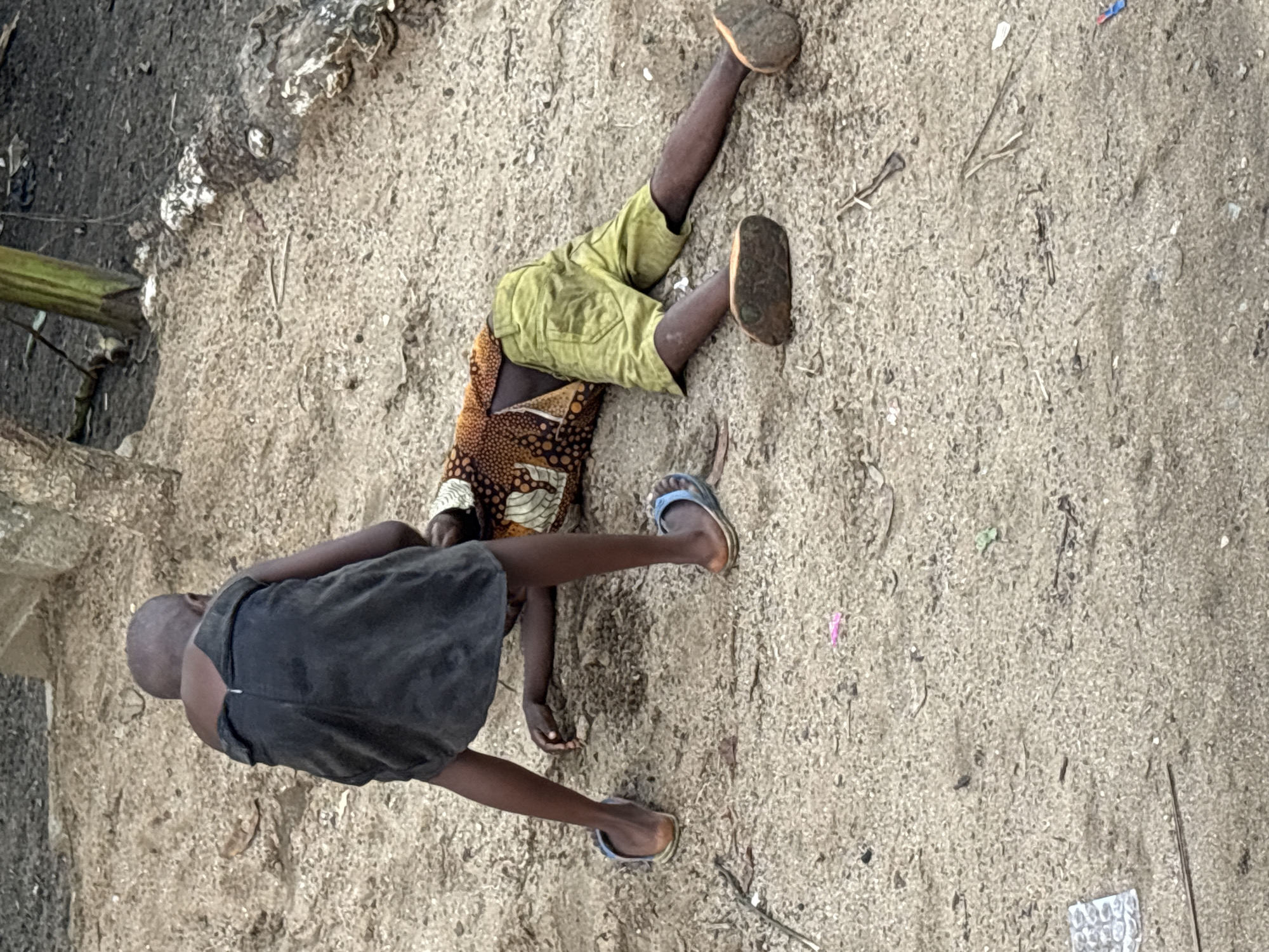 Two children lie on a sandy surface adjacent to a building in Ikot Ekpene, Nigeria. One child wears a black t-shirt and blue sandals, and the other wears a patterned brown and white top, yellow shorts, and brown sandals. The children are interacting with each other, their bodies touching. The sandy surface contains scattered debris, including sticks, leaves, and a foil blister pack. A building is visible to the left of the frame, with a small section of wall and a wooden post.