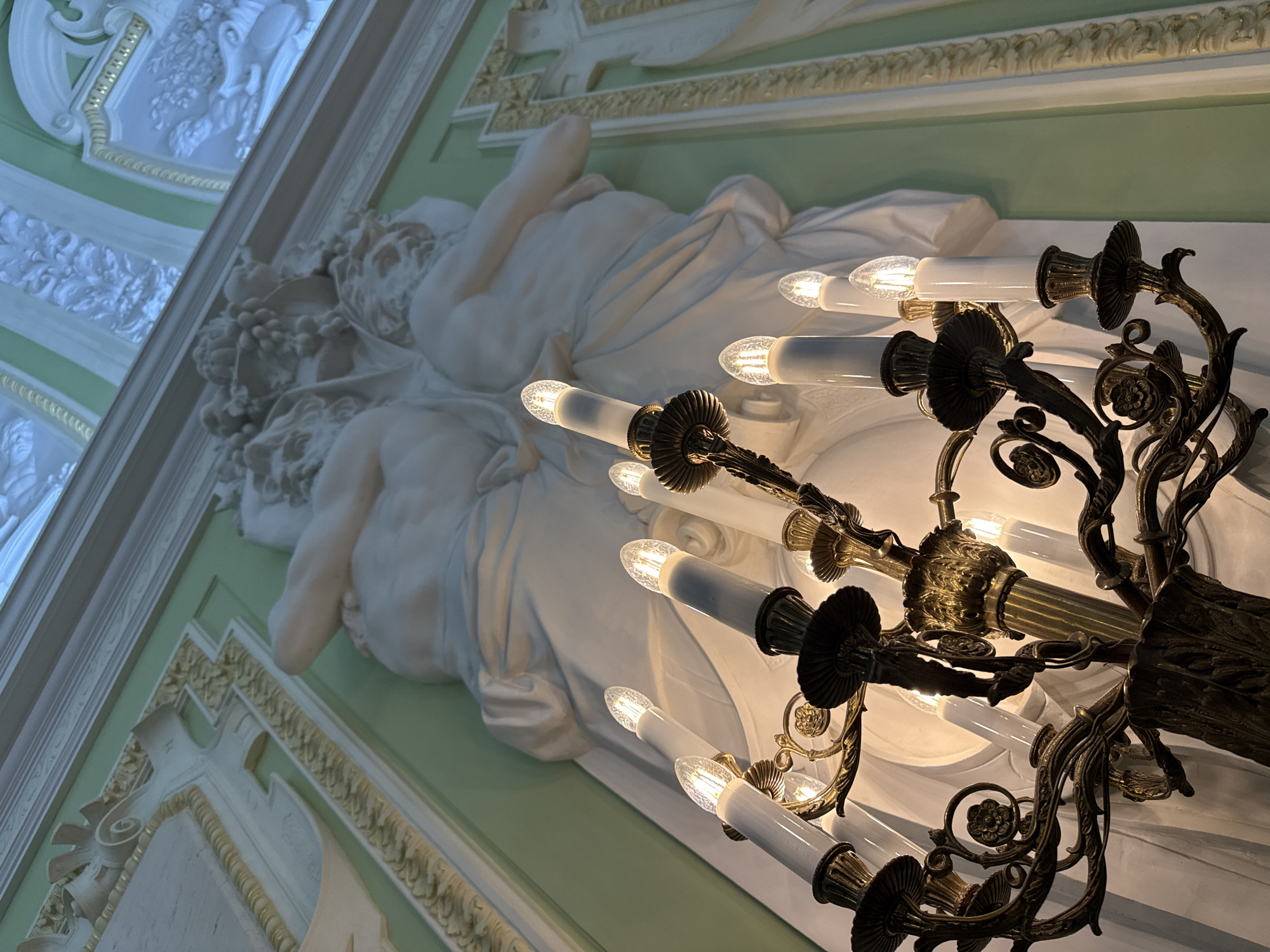 The image shows a close-up view of an ornate brass chandelier with faux candle lights, positioned in front of a sculpted plaster frieze and a light green wall. The frieze depicts what appears to be two cherubic figures intertwined, adorned with grapes. The chandelier has multiple arms with detailed scrollwork and floral accents, each holding a white, faux candle with a visible filament. The overall aesthetic suggests an interior of a historic or opulent building. Without additional context or identifying landmarks, the specific location in Nizhny Novgorod, Russia cannot be determined.