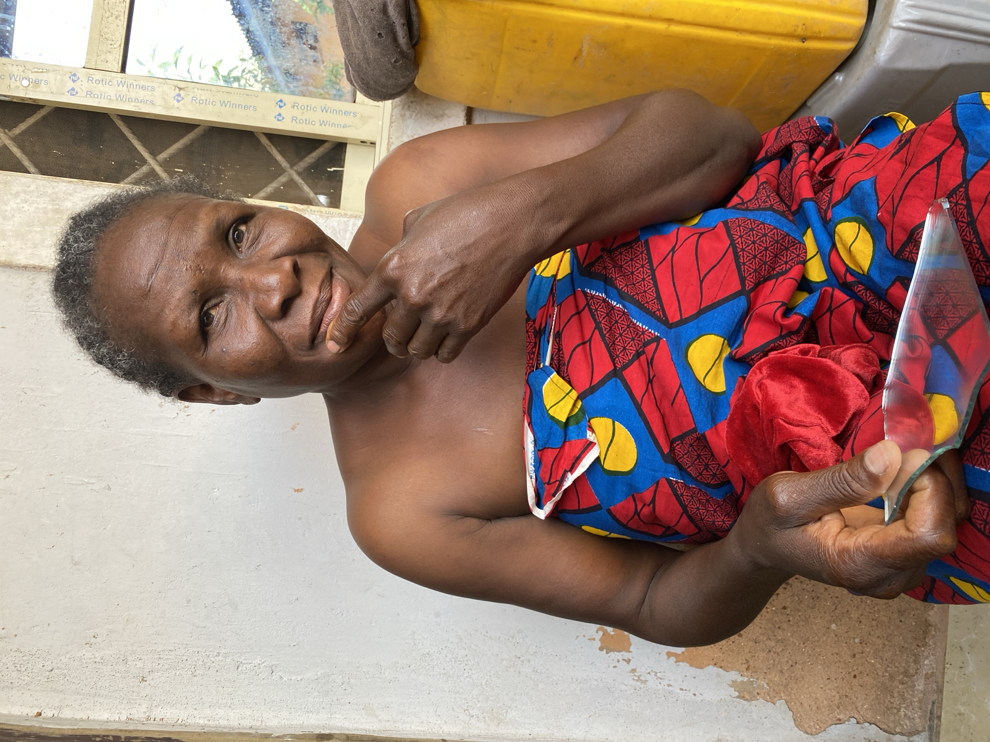 An older African woman is depicted in a close-up shot. She has short, graying hair and dark skin. She is topless, with a red, blue, and yellow patterned cloth draped around her lower body. In her right hand, she holds a piece of broken glass. Her left hand is raised near her face, with a finger pointing to her chin. The background includes a white wall, a window, and a yellow container. The location is Bukuru, Nigeria.