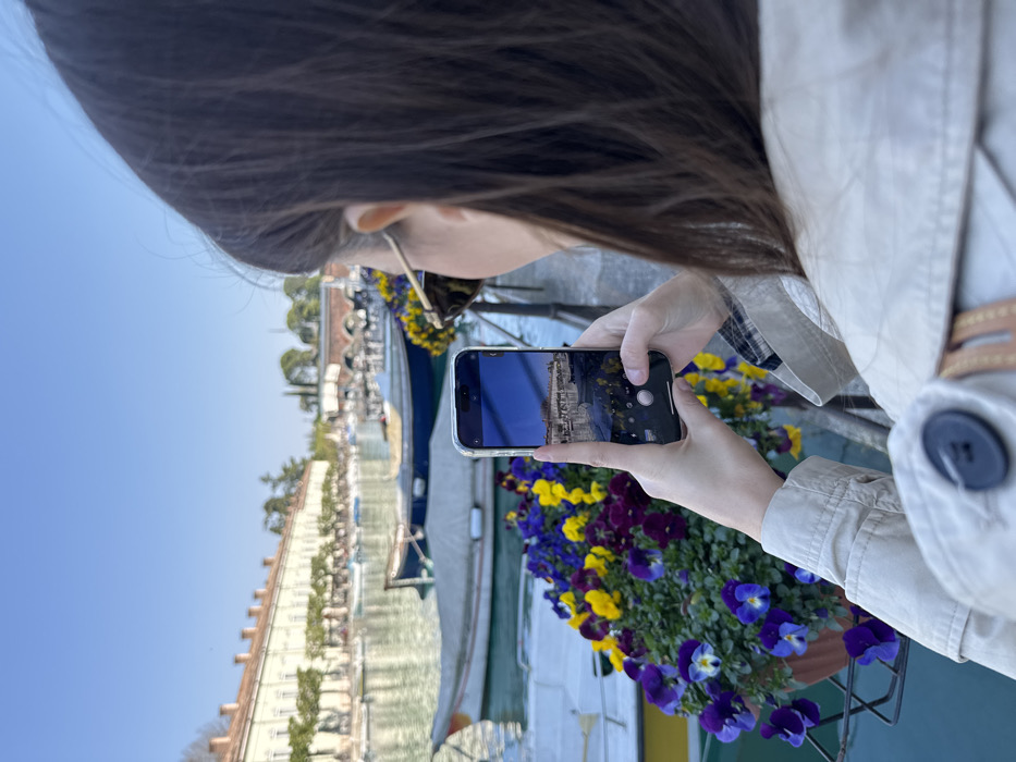 A person with brown hair, wearing sunglasses and a light-colored coat, stands with their back mostly to the camera, facing a canal in Peschiera del Garda, Italy. The individual holds a silver-colored smartphone horizontally with both hands, actively photographing or recording the scene, which is displayed on the phone's screen. In the immediate foreground to the left, a planter overflowing with vibrant purple, yellow, and blue pansies is visible. The midground features calm, light-blue water populated by multiple small boats, some covered with light-toned tarpaulins. The background is dominated by a long, multi-story, light-colored historic building with numerous uniform arched openings (Caserma d'Artiglieria) lining the far bank of the canal. Further in the distance, a multi-arched brick bridge spans the water. The sky above is clear and blue.