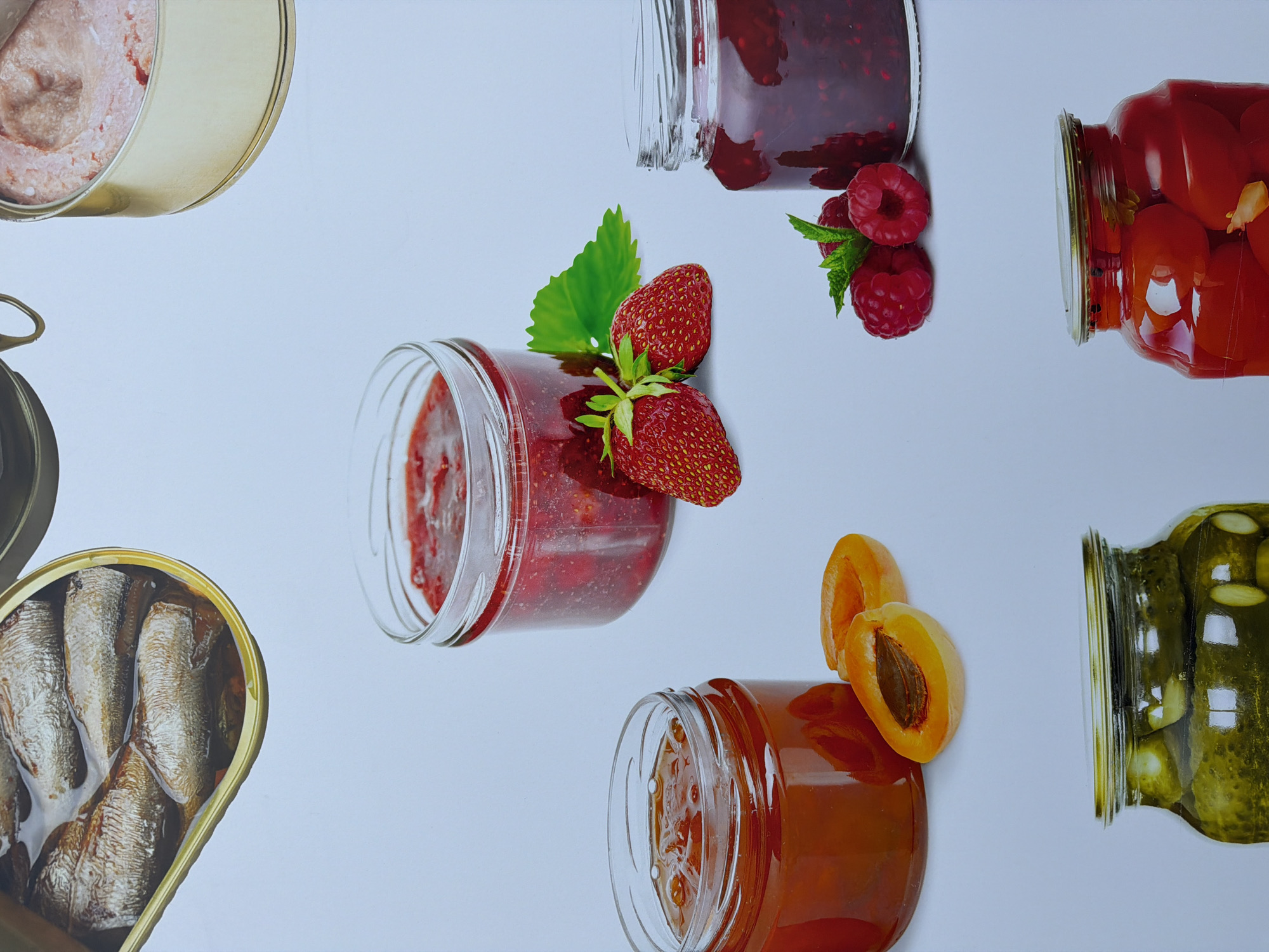 The image shows a flat lay arrangement of various preserved foods on a light blue surface. The items include canned fish (sardines), pate, raspberry jam with fresh raspberries, strawberry jam with fresh strawberries, apricot jam with fresh apricot halves, pickled tomatoes, and pickled cucumbers. The background is a uniform light blue. The scene does not contain any discernible characteristics or landmarks that would allow for the determination of its location in the city of Chicago, United States of America, or anywhere else. There are no people present in the image.