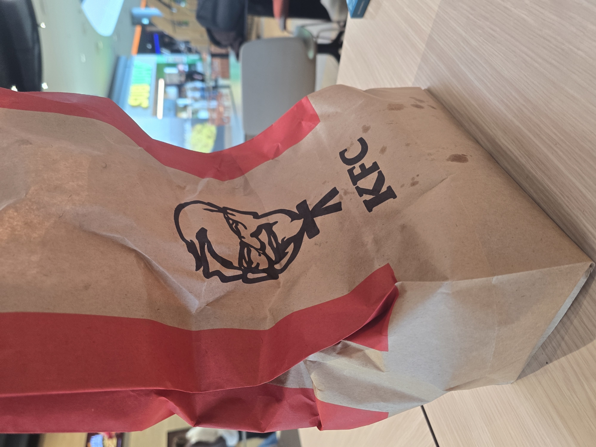 The image shows a paper bag from KFC resting on a light-colored wooden surface. The KFC logo and lettering are visible on the bag. The bag is predominantly tan, with a red band running along the top. In the background, there's a blurred view of what appears to be a food court or shopping center, potentially including signage for Subway. Based on the visual clues, the setting might be within a mall or food court. The location is not explicitly stated in the image, so the city of Burgas, Bulgaria cannot be confirmed.