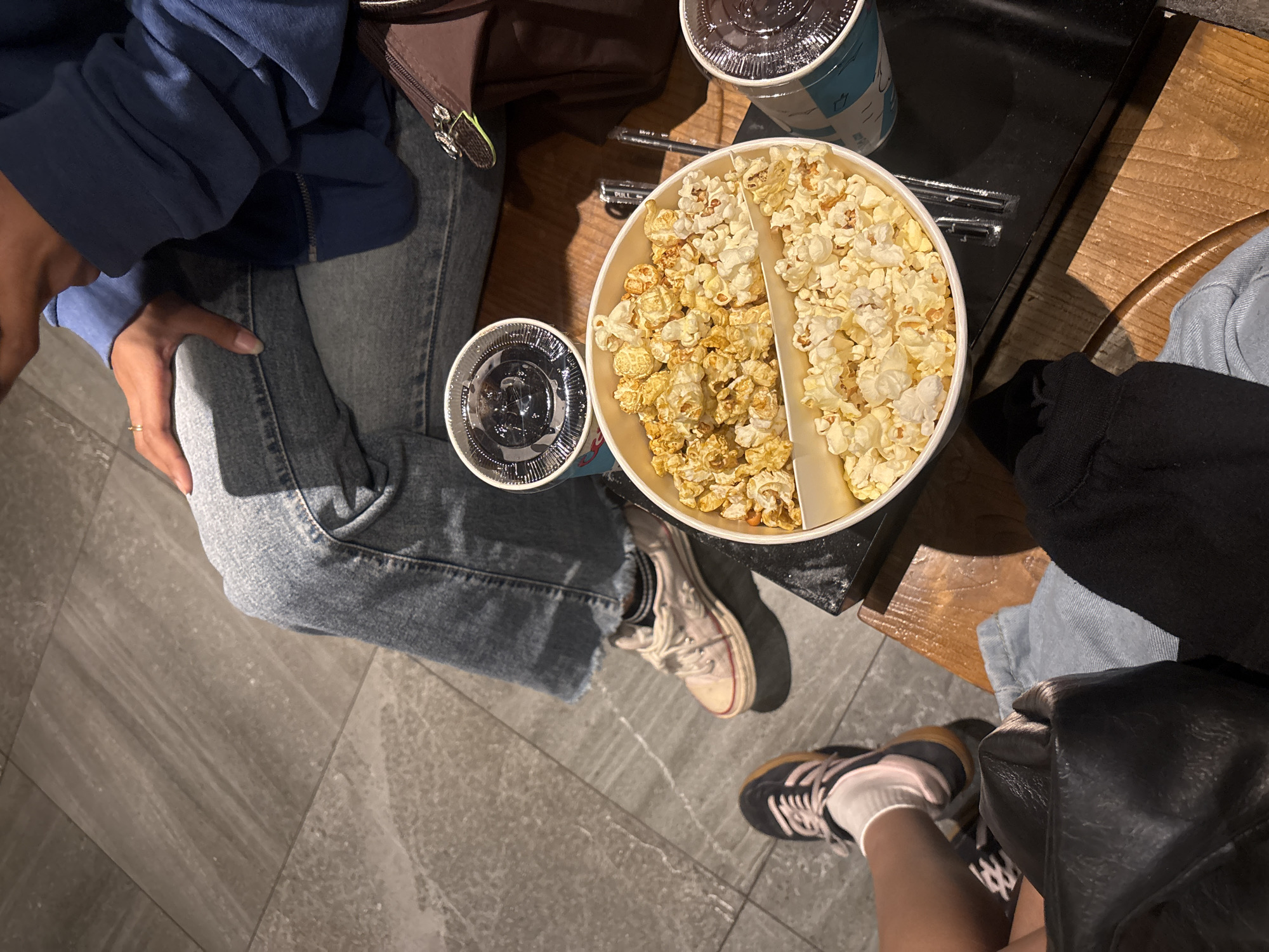 The image shows a top-down view of two people sitting, likely in a cinema or entertainment venue. The floor is tiled with large, grey, rectangular tiles. In the foreground, a white container divided into two sections holds popcorn, with one section appearing to be a caramel or buttered variety and the other plain. Two cups with clear lids and dark beverages are placed near the popcorn.
To the left, a person's legs are visible, wearing dark sneakers with white socks and light blue jeans. A black leather bag and a black garment are also in the lower left corner.
To the right, another person's legs are visible, wearing light-colored sneakers with dark stripes and light blue jeans. This person is wearing a blue hoodie, and their hand, adorned with a ring, rests on their leg. A brown bag is situated to the right of this individual.
The scene suggests a casual outing, possibly a movie date, in a public space with a modern aesthetic. The presence of popcorn and beverages indicates a leisure activity. The overall lighting is dim, typical of a cinema. The cropped images offer a closer look at the textures of the floor tiles, the clothing details, and the popcorn container. The context of West Jakarta, Indonesia, adds a geographical element to the setting.