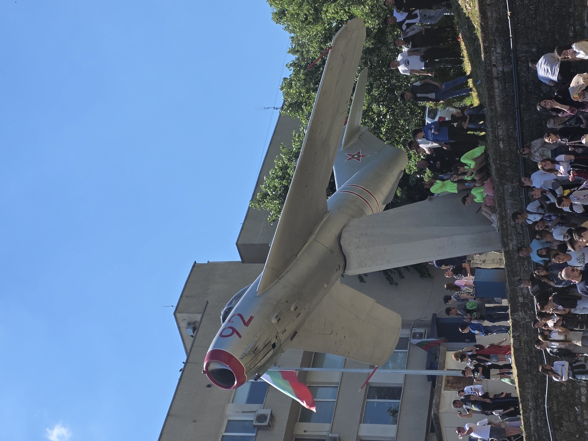 The image depicts a MiG-17 fighter jet mounted as a monument on the exterior of a building in Yambol, Bulgaria. The aircraft is painted silver with red accents, including the nose cone and the number "92" on the fuselage. A red star is visible on the vertical stabilizer. A Bulgarian flag is draped near the jet's nose. A crowd of people is gathered along a wall, likely observing or participating in an event related to the monument. The backdrop includes a building with visible windows and an air conditioning unit. The sky is clear and blue. Trees are visible in the background. The image appears to be taken during the day.