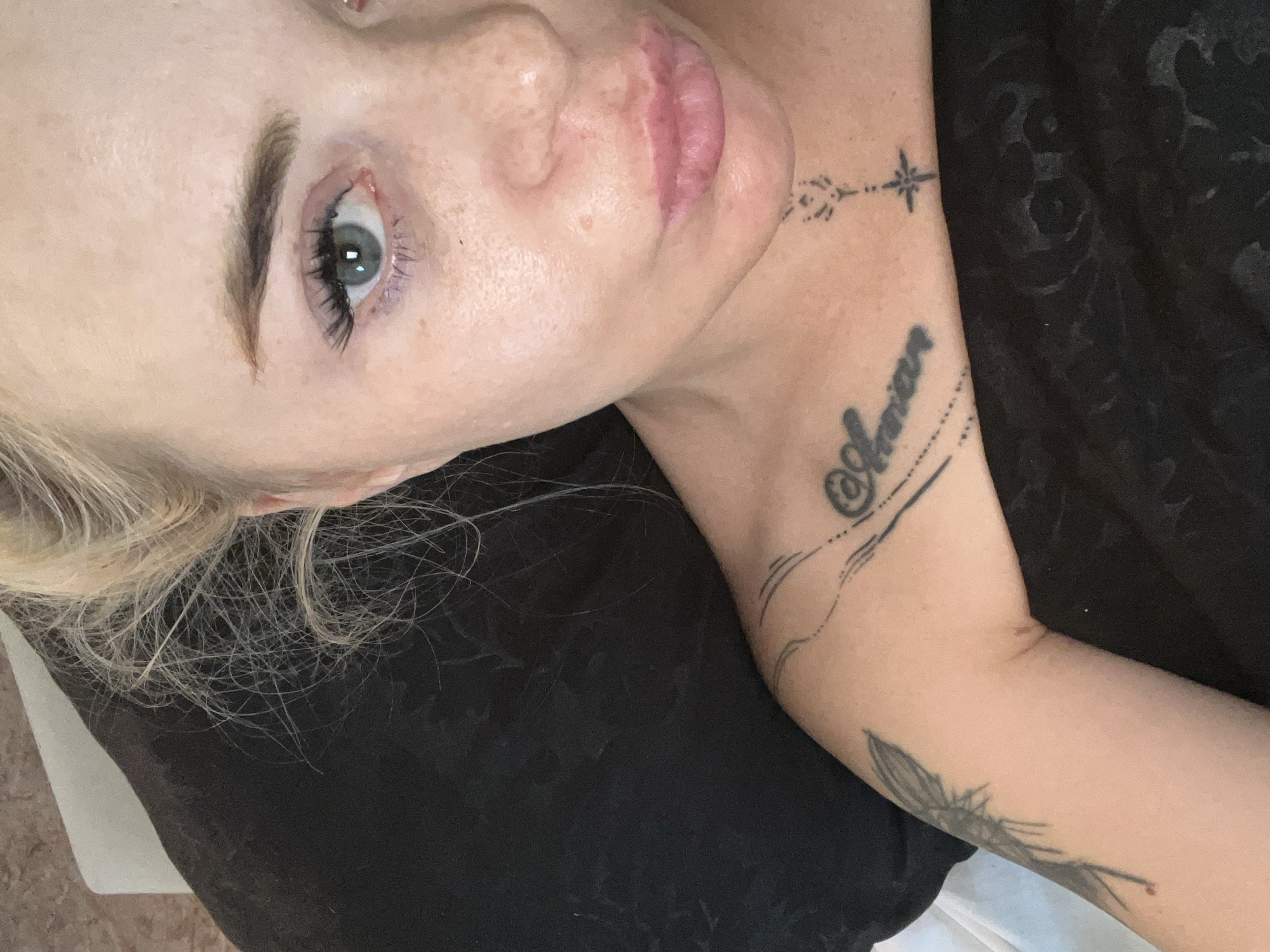 This media file is a close-up, high-angle shot of a person's face and arm. The person has blonde hair and blue eyes, and is wearing makeup. They have several tattoos visible on their arm and neck. One tattoo on the neck appears to be a star with a line extending from it, and another on the arm reads "Amour" in cursive. There is also a feather tattoo on the forearm. The background is dark and appears to be clothing. The lighting suggests an indoor setting. The person's expression is neutral, and the overall mood is casual. No specific activity or event is clearly depicted.