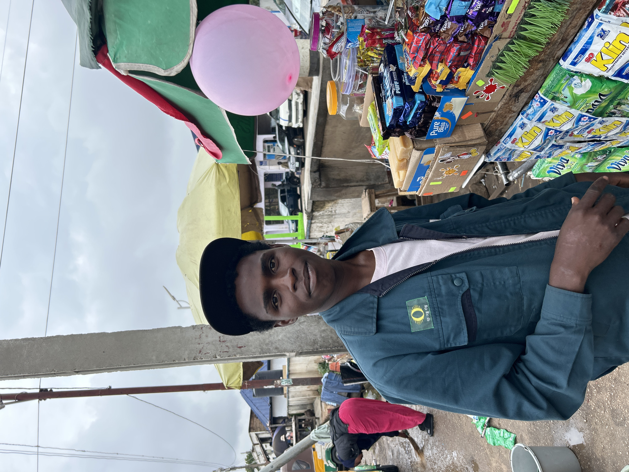 The image captures a street scene in Jos, Nigeria, under an overcast sky. A young Black man, wearing a dark teal jacket with an insignia on the left breast and a black baseball cap, stands facing the camera. He has short, dark, curly hair and is looking directly at the viewer. The jacket is zipped up, revealing a light-colored t-shirt underneath.
To the right of the man, a stall is laden with various goods. These include stacks of packaged snacks and toiletries, such as boxes of "Puff Wafers," "Pure Bliss" biscuits, and multiple packets of laundry detergent, including brands like "Viva Plus" and "So Klin." A large pink balloon is tied to the top of the stall's awning, which is a combination of green and red fabric. Glass jars filled with small items are also visible on the stall.
In the background, to the left of the man, another person, dressed in a pink outfit, is sweeping the wet ground with a broom. Behind them, a small yellow awning covers part of a building, and further back, several parked vehicles, including cars and a tricycle, are visible. The ground appears wet, suggesting recent rain. Power lines and a concrete pole are prominent elements in the background. The overall impression is that of a bustling street market or roadside stall.