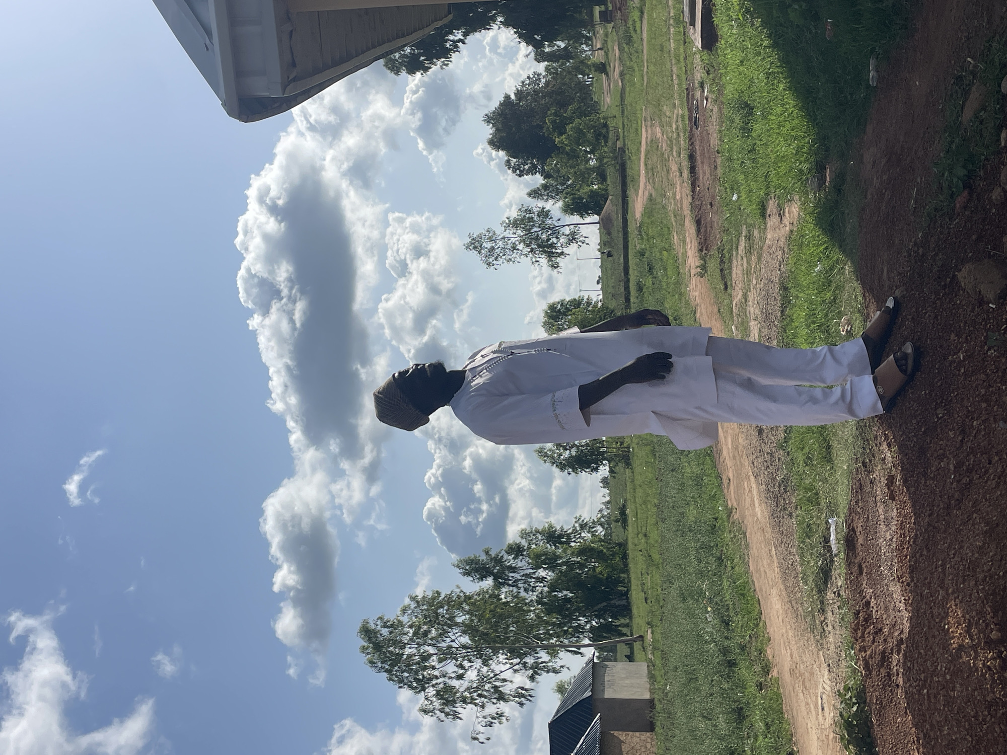 A man wearing all white clothing is standing on a dirt path in Durumi, Nigeria. The path is lined with green grass and trees. The sky is blue and cloudy. A building is visible in the background.
