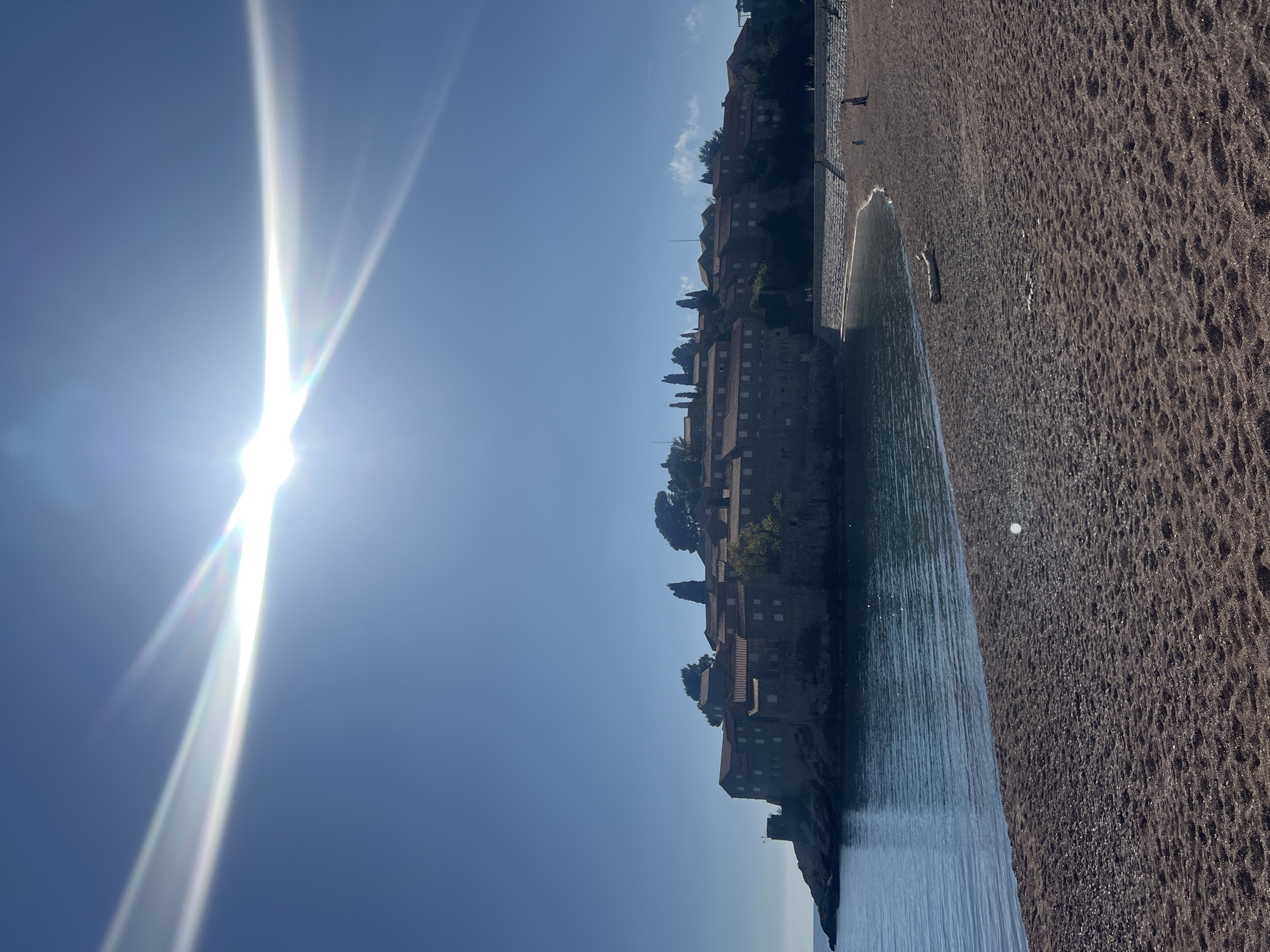 The image shows a sunny view in Sveti Stefan, Montenegro. The sun shines brightly in the blue sky, creating a lens flare effect. The foreground is dominated by a beach with dark sand. The sea is adjacent to the beach, and the water appears calm. The iconic island of Sveti Stefan, with its cluster of buildings, is visible in the middle ground. The buildings appear to be old and made of stone, with red tiled roofs. Some greenery, possibly trees or bushes, is also visible amongst the buildings. Several people are walking along the beach, though they appear small in the distance.