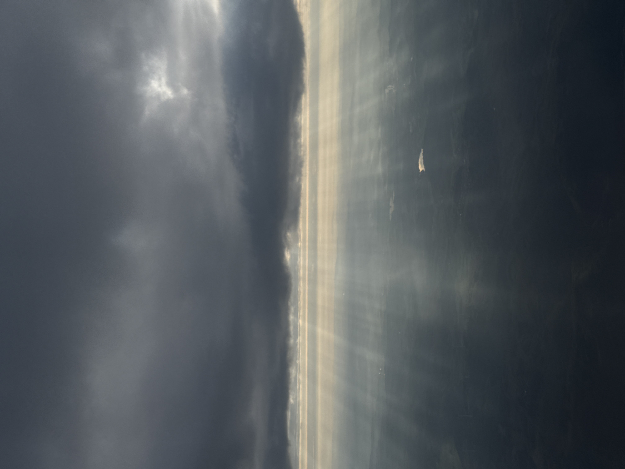The image depicts a view of a cloudy sky with sunbeams breaking through. The left side of the image features dark, heavy clouds. Between the clouds and a dark area on the right, there are beams of sunlight streaming downward. The image appears to be taken from an elevated position. It is not possible to determine the location in the city of Freudenburg, Germany from this image.