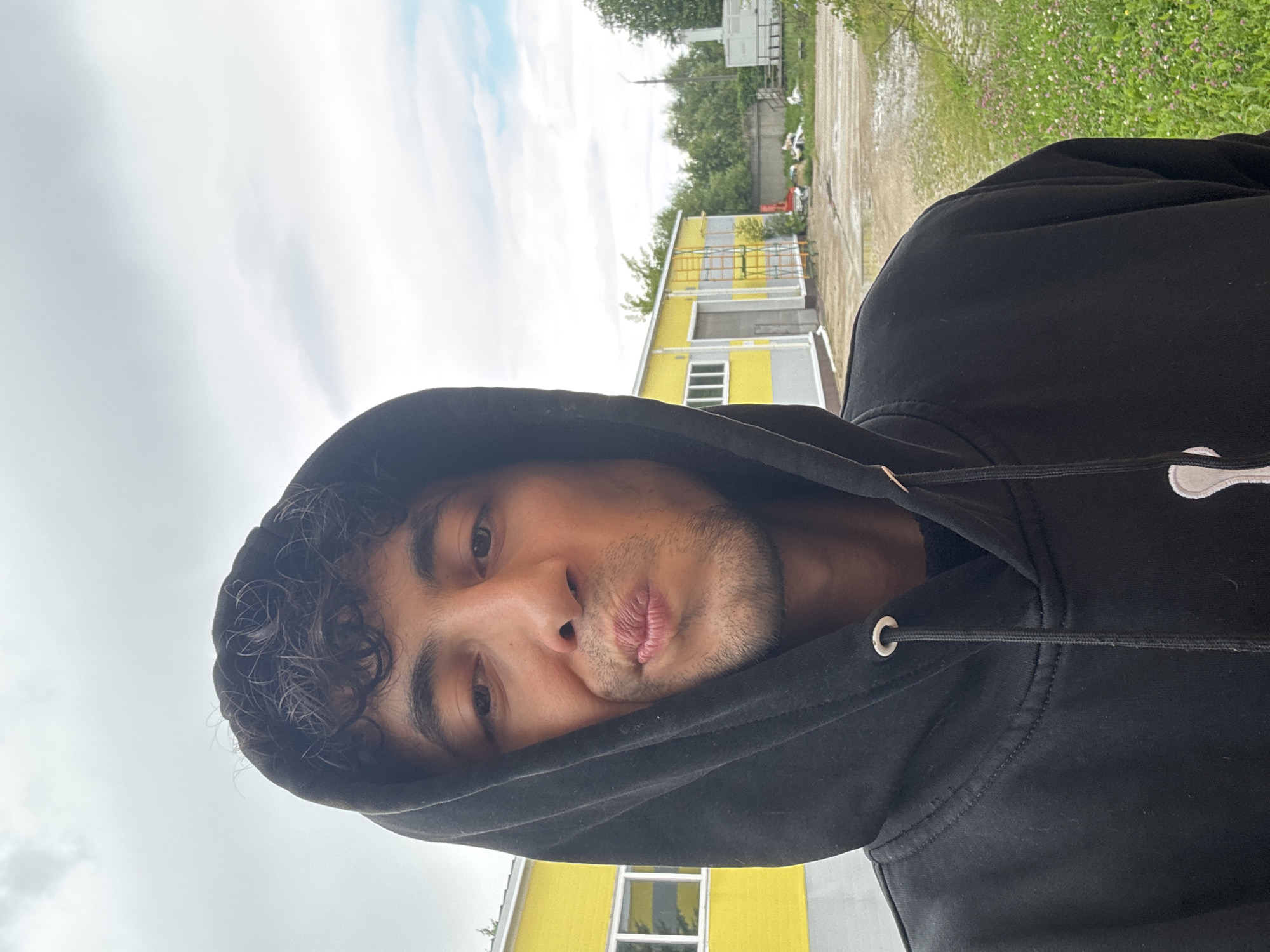 A close-up, selfie-style photo shows a young man wearing a black hooded sweatshirt, pulled up to frame his face. He has dark, curly hair and is making a kissing face. His eyes are looking directly at the camera. In the background, a yellow and grey building with a metal garage door is visible, along with some greenery and a cloudy sky. The ground appears wet, with puddles and a grassy area dotted with small flowers.
