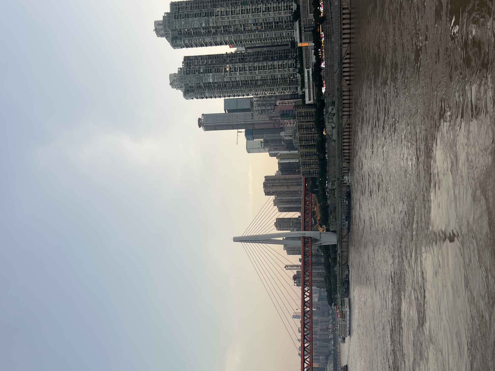 The image captures a wide shot of the Yangtze River in Nan'an District, Chongqing, China, under a hazy sky. In the foreground, the murky brown waters of the river occupy the bottom half of the frame. Several boats, including a larger passenger vessel with a white hull and blue accents, are visible on the water.
Dominating the middle ground is the Hongyadong Bridge, a distinctive red suspension bridge. Its central white pylon stands tall, supporting the bridge's cables. The bridge spans across the river, connecting the Nan'an District to the opposite bank, which is lined with a dense cluster of high-rise buildings.
The background showcases the Chongqing skyline, characterized by numerous modern skyscrapers of varying heights and architectural styles. The buildings appear to be residential or commercial towers, with many featuring reflective glass facades. The overall impression is that of a bustling urban environment situated along a major river. The time of day appears to be late afternoon or early evening, given the soft lighting and the pale sky.