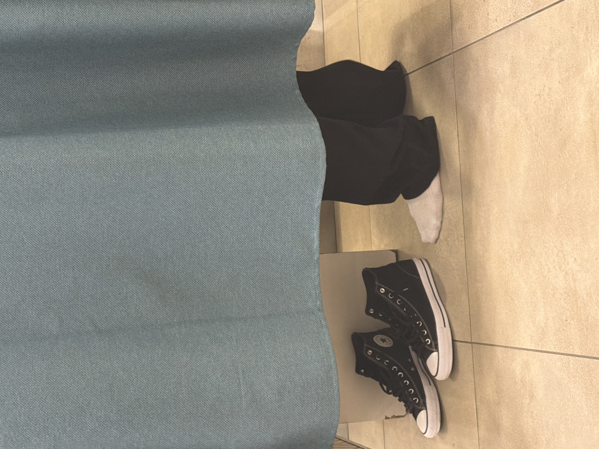 A person wearing black pants and a white sock sits behind a blue curtain, with a pair of black high-top Converse sneakers beside them on a tile floor. No information indicates the location is Moscow, Russia.