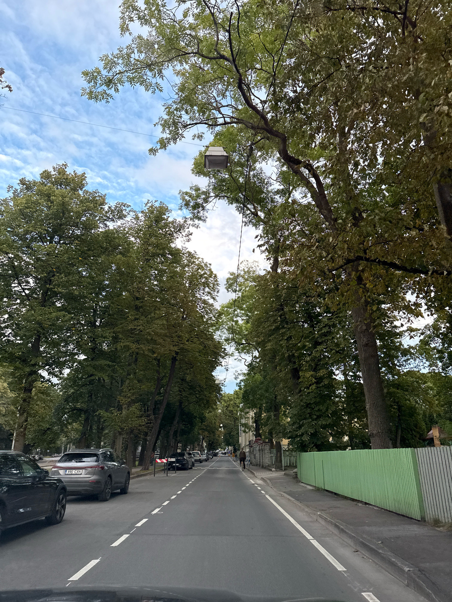 A view from a vehicle driving down a tree-lined street in Tallinn, Estonia. The road is paved and has lane markings. On the right side of the road, there is a sidewalk and a light green corrugated metal fence. On the left side of the road, there are parked cars and more trees. The sky is blue with some white clouds. In the distance, there are buildings and people walking and cycling. A box-like object is hanging from a wire between two trees. The license plate of the grey car in the foreground on the left is 890 CXN.