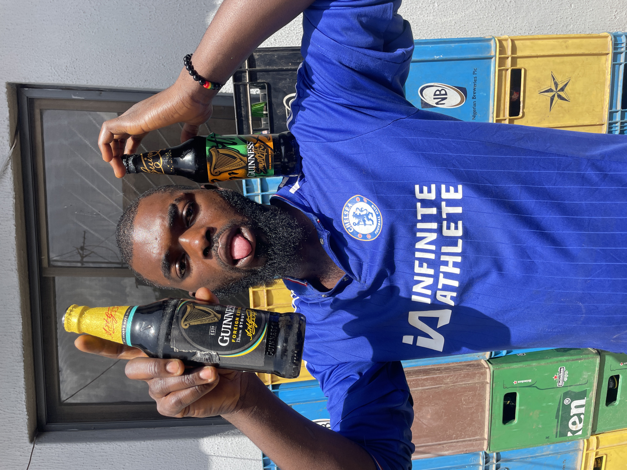 A young Black man with a beard and a blue Chelsea FC jersey is standing in what appears to be an outdoor storage area. He is holding two bottles of Guinness beer, one in each hand, above his head. The man is sticking his tongue out. The background consists of stacks of colorful plastic crates, some of which are labeled with brewery logos. The lighting suggests it is daytime, likely under direct sunlight. The overall environment is utilitarian and appears to be in an urban setting within Jos, Nigeria, indicated by the branding on the crates.