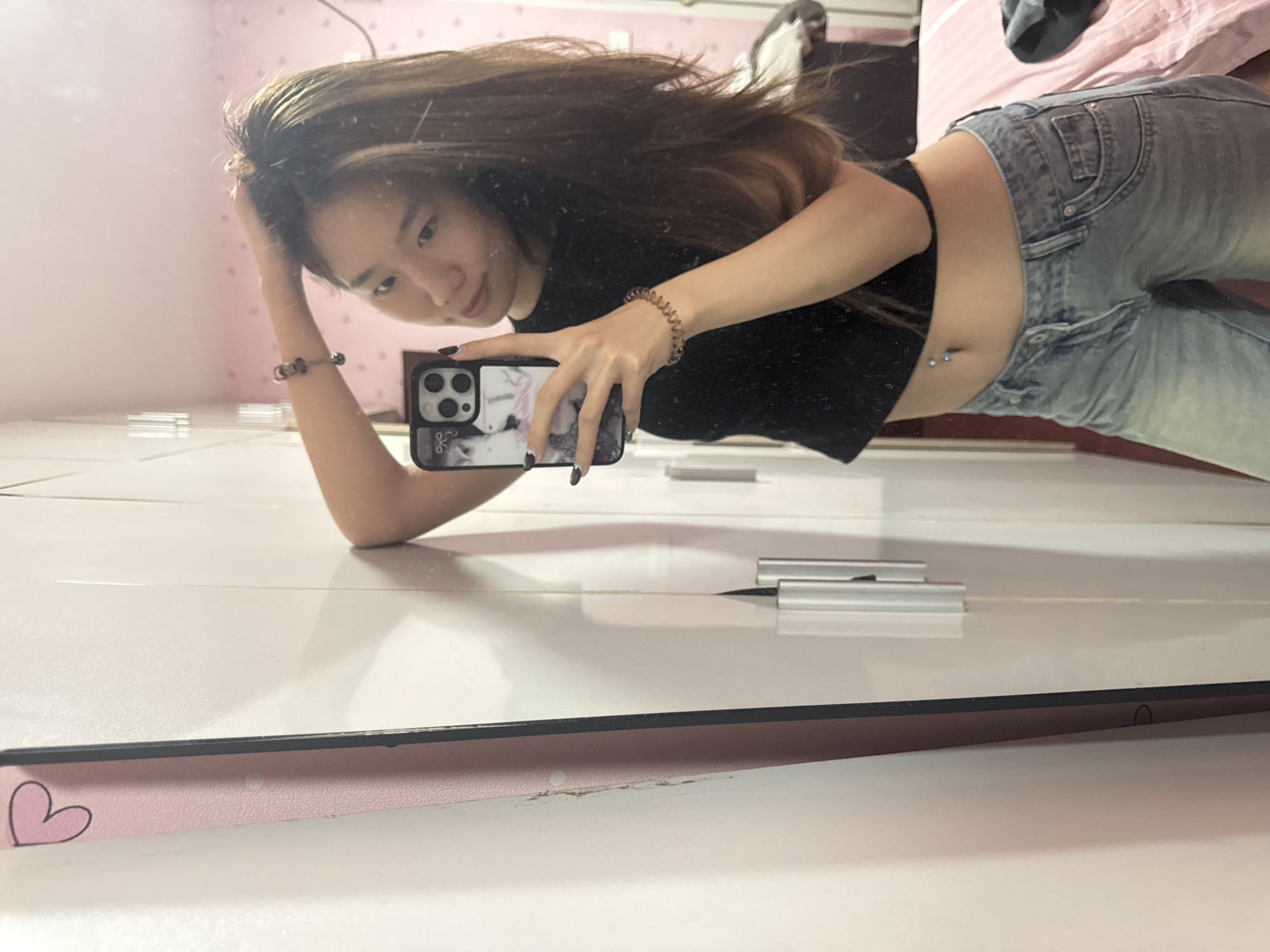 A young Asian woman is posing for a selfie in a mirror. She is wearing a black crop top and jeans. Her hair is long and brown. She is holding a smartphone with a decorated case. The background appears to be a bedroom with pink walls and furniture. The mirror has a pink frame with a heart design. It is impossible to determine the location of Shapingba District, China from this image alone.