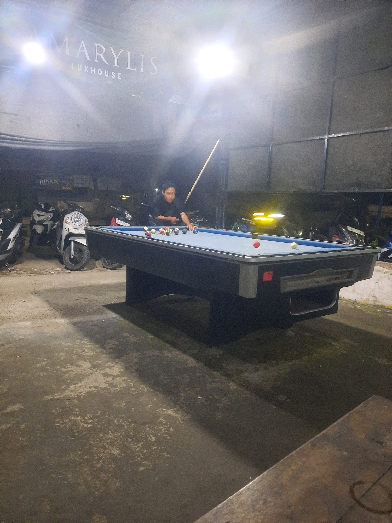 A billiard table is set up outdoors in Pucangtelu, Indonesia, under a banner displaying "MARYLIS LUXHOUSE." A young person is seen playing billiards, holding a cue stick and looking at the balls on the table. Several motorcycles are parked behind the billiard table, and the ground appears to be a concrete surface. The lighting suggests it is nighttime, with bright spotlights illuminating the scene.