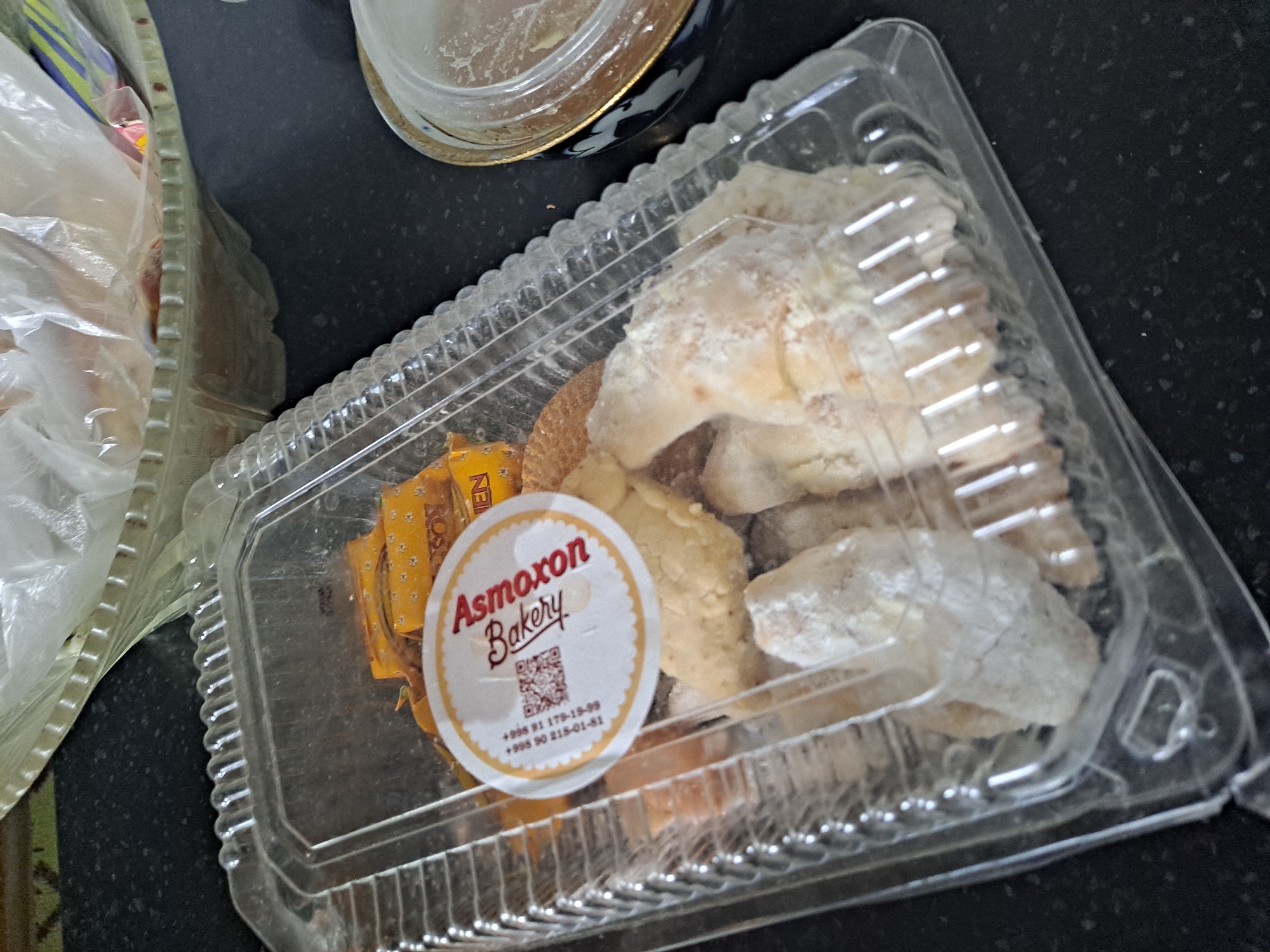 The image shows a clear plastic container filled with baked goods, likely cookies or pastries, some of which are dusted with powdered sugar. A circular sticker with the text "Asmoxon Bakery" and a QR code is affixed to the lid. The container is placed on a black speckled countertop. In the background, a part of a green woven basket with a white plastic liner is visible, suggesting the container might be within a larger shopping bag or picnic basket. The overall scene appears to be a domestic or retail setting, likely indoors. The location is identified as Namangan City, Uzbekistan.