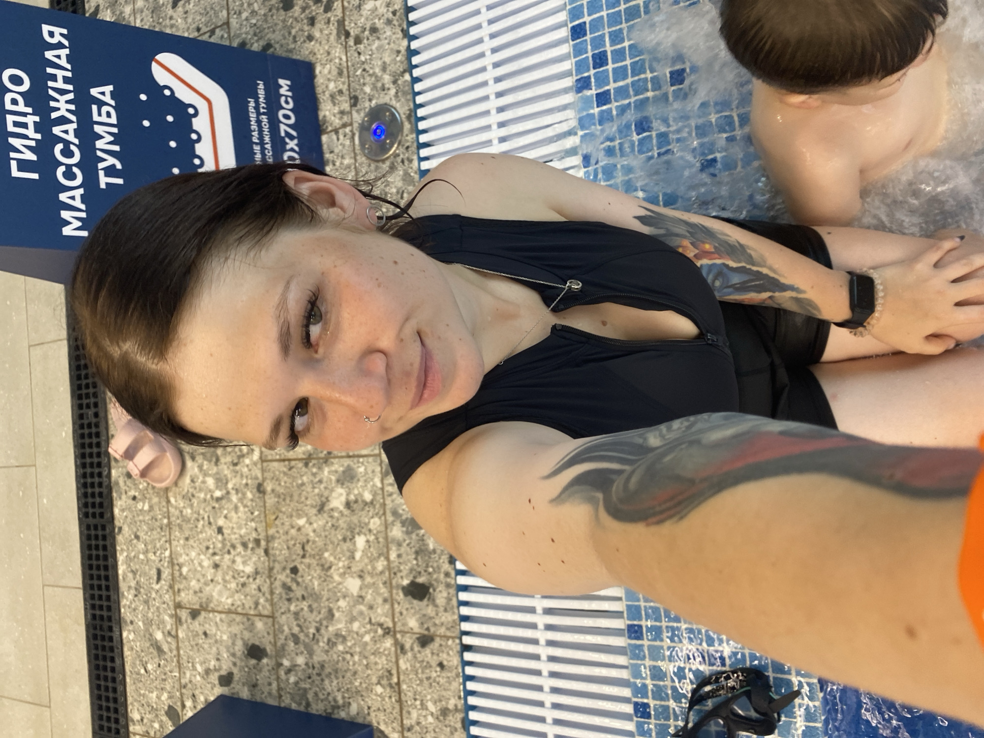 The image shows a woman taking a selfie in a jacuzzi. She is wearing a black swimsuit and has tattoos on her arms. A child is visible in the water beside her. In the background, there is a sign in Russian that says "Hydro Massage Bath." The floor is tiled, and there are pink sandals on the floor. The image appears to have been taken in Votkinsk, Russia.