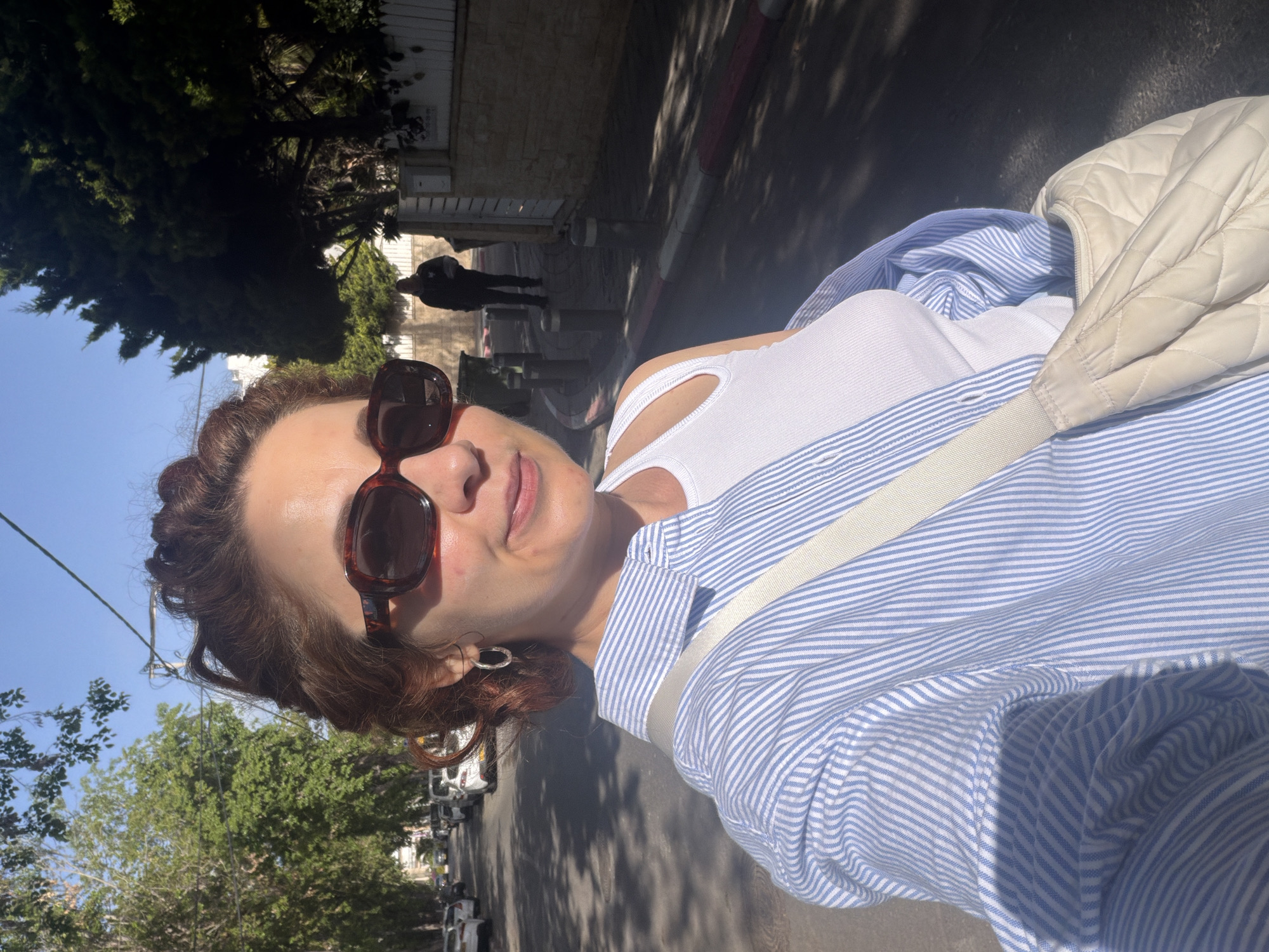 A woman is taking a selfie on a sunny day in Bat Yam, Israel. She is wearing sunglasses, a white tank top, and a blue striped shirt, with a quilted white bag over her shoulder. In the background, there's a street with parked cars, trees, and a building. A person is walking in the distance.