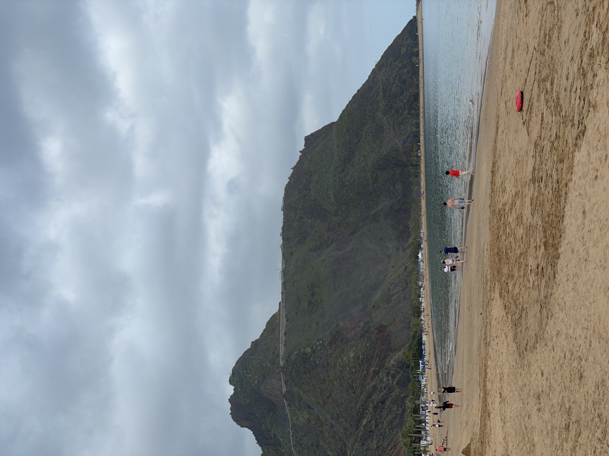 The image displays a panoramic view of a coastal scene under an overcast sky. A large, dark, and rugged mountain, covered in vegetation, dominates the left side of the frame, descending towards the ocean. The calm, grey-blue water of the sea stretches from the base of the mountain towards the horizon on the right.
In the foreground, a wide expanse of light brown sandy beach occupies the right side of the image. Several people are scattered along the beach and in the shallow water. Some individuals are seen walking along the shore, while others are wading or standing near the water's edge. A bright red bodyboard or float rests on the sand in the mid-foreground. Further down the beach, rows of sun loungers and umbrellas indicate a developed beach area.
The weather is cloudy, with heavy grey clouds covering the sky, suggesting an overcast daytime. Despite the subdued lighting, people are out enjoying the beach. The scene depicts a serene yet active beach environment, characteristic of coastal regions. Based on the provided context, the location is Santa Cruz de Tenerife, Spain. No visible text is present in the image.