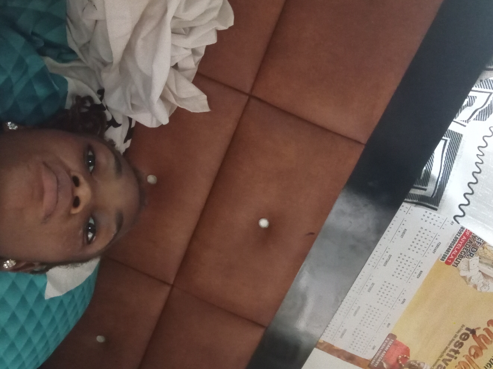A close-up, slightly rotated, indoor shot shows a woman lying down next to a calendar. The woman's face, turned toward the camera, is partially covered by a white and blue patterned cloth. She wears what appears to be earrings. Beside her, a brown cushioned surface with white button accents is visible. To the right of the cushioned area is a table or surface covered by a black strip and a calendar. Text on the calendar is partially readable and includes the names of months (February, March, April, May, August, September) and the phrase "EF DR. YADEGBUYI OF AIYELALA 1". The location is in AIYELALA 1.