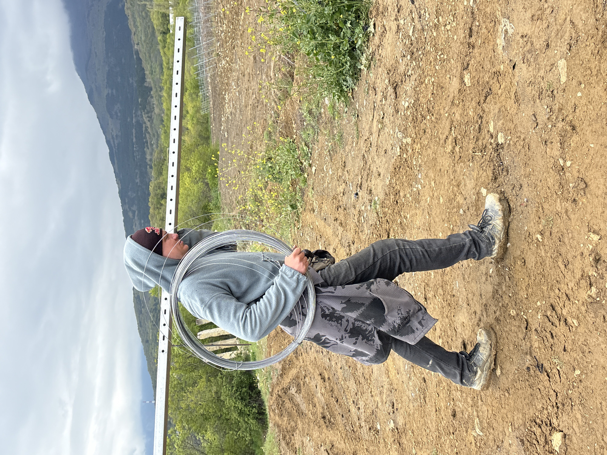 A man stands on a dirt field in an outdoor setting. He is wearing a gray hooded sweatshirt, dark pants, and muddy shoes. He is also wearing a baseball cap and has a gray and black patterned garment tied around his waist. He is holding a large coil of metal wire in front of him. Behind him, there is a white fence-like structure, green foliage, and a mountain in the distance under a cloudy sky. The location is not identifiable as Pushkino, Ukraine based on visual information.